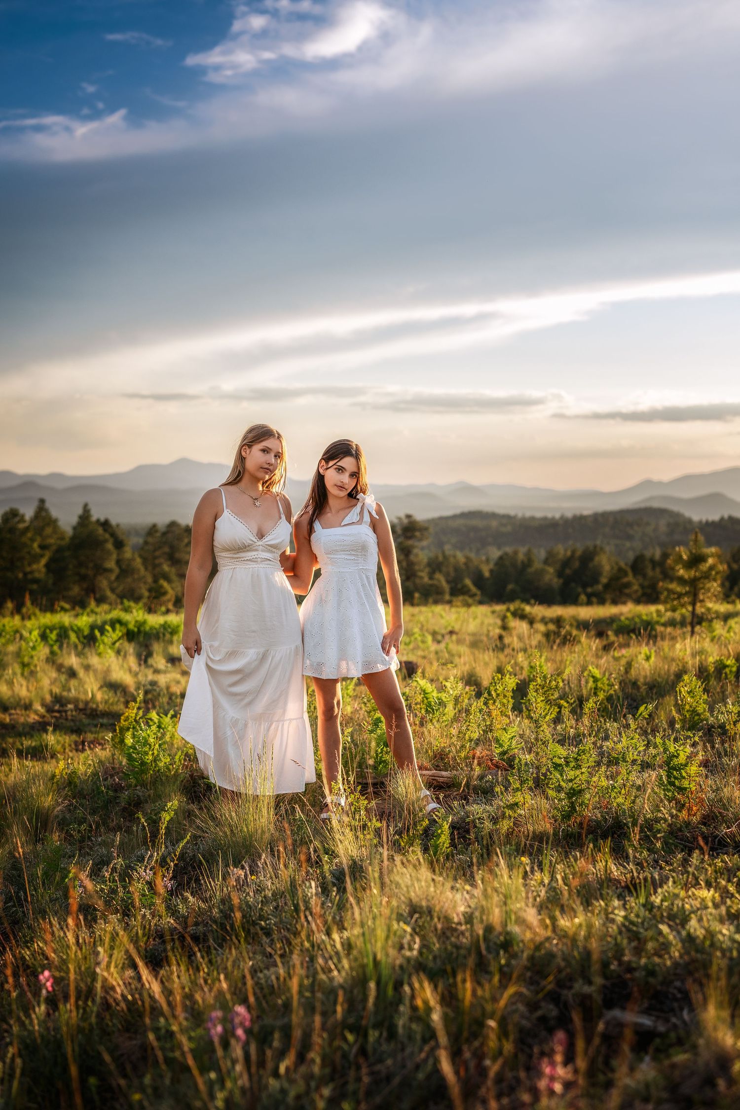 Two girls in white dresses holding hands in a scenic mountain meadow at golden hour.