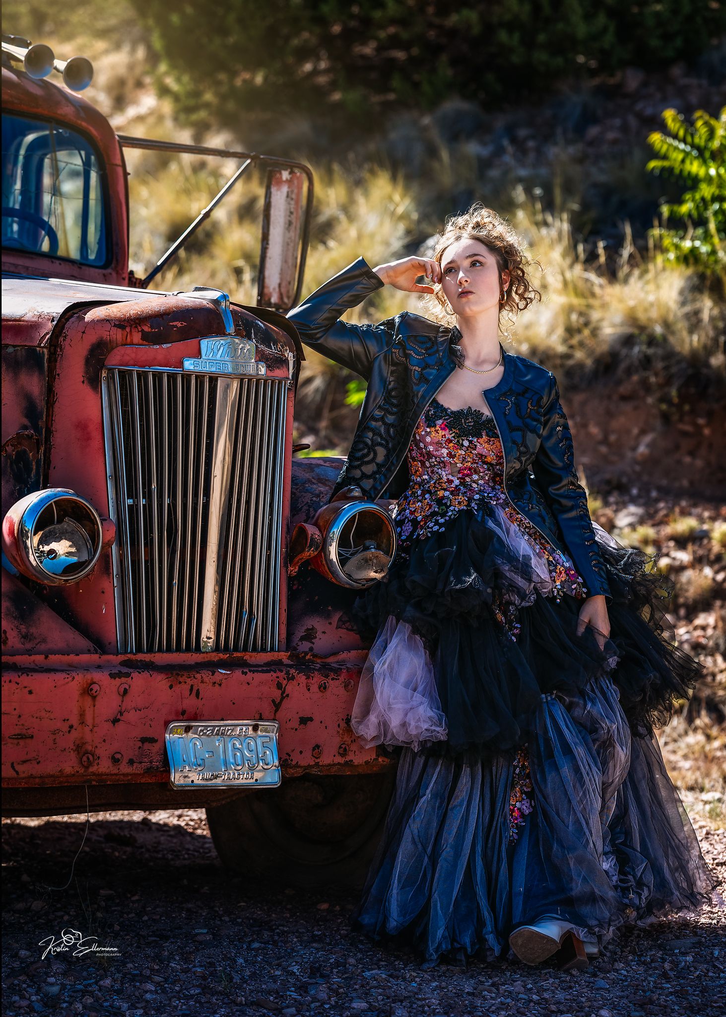 Woman in floral gown and leather jacket poses beside a vintage red truck on a rural dirt road.