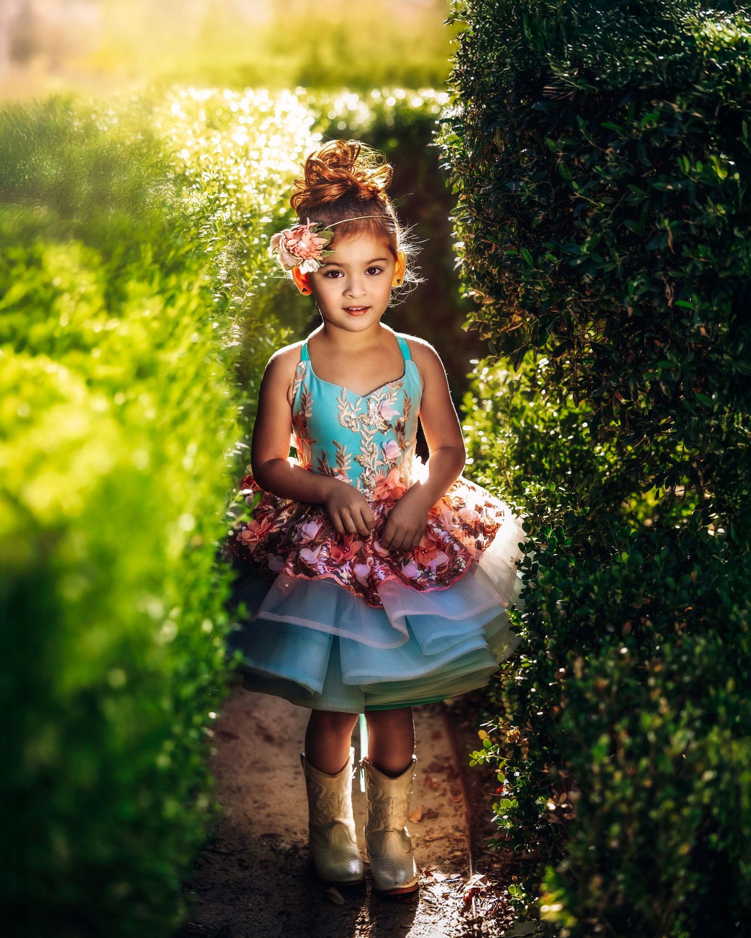A young girl in a colorful tiered tutu dress and boots walks through a lush green garden path.