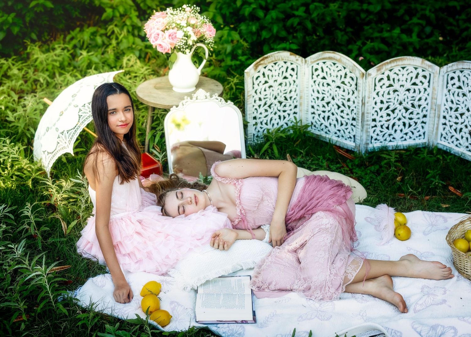 Two girls in pink dresses enjoy a garden picnic, one reading while the other rests on a white blanket.