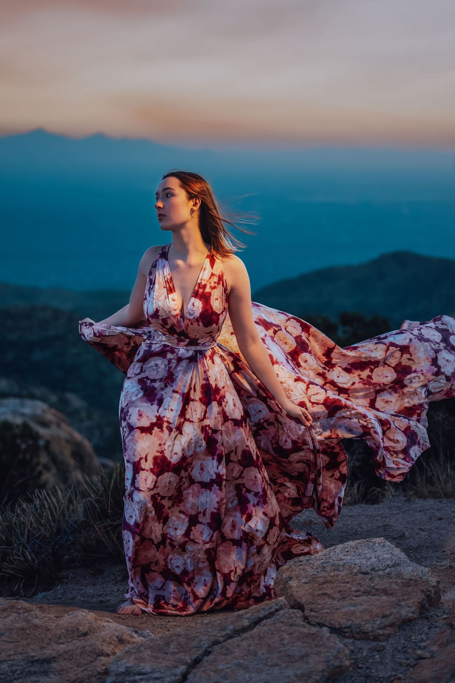 Woman in billowing floral maxi dress stands on rocky summit with mountain vista at dusk.