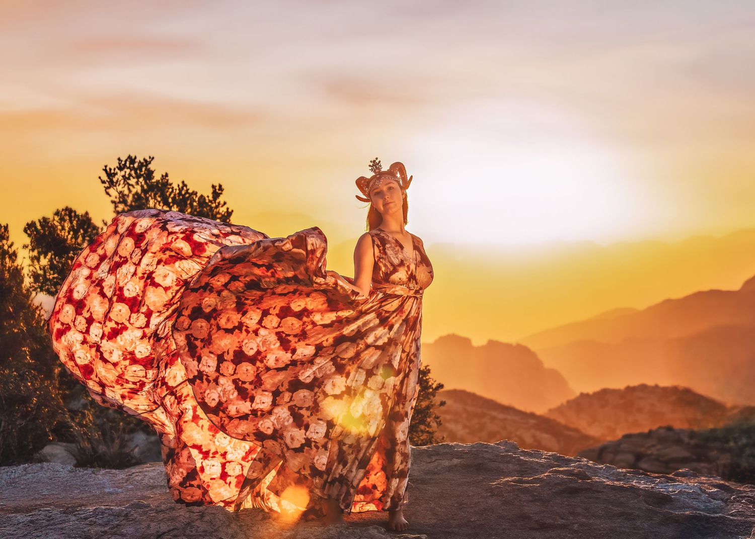 Woman in flowing floral dress dancing on mountain rocks with warm golden sunset behind her.
