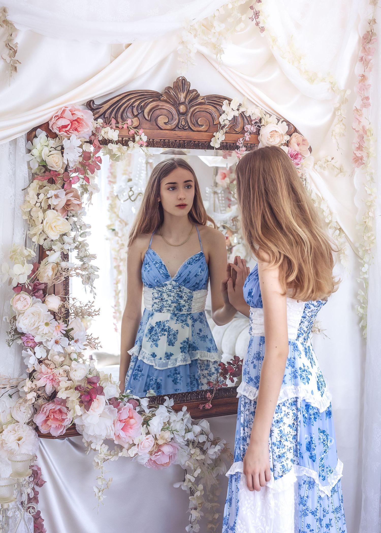 A young woman in a blue floral dress gazes into an ornate mirror surrounded by fresh flowers.