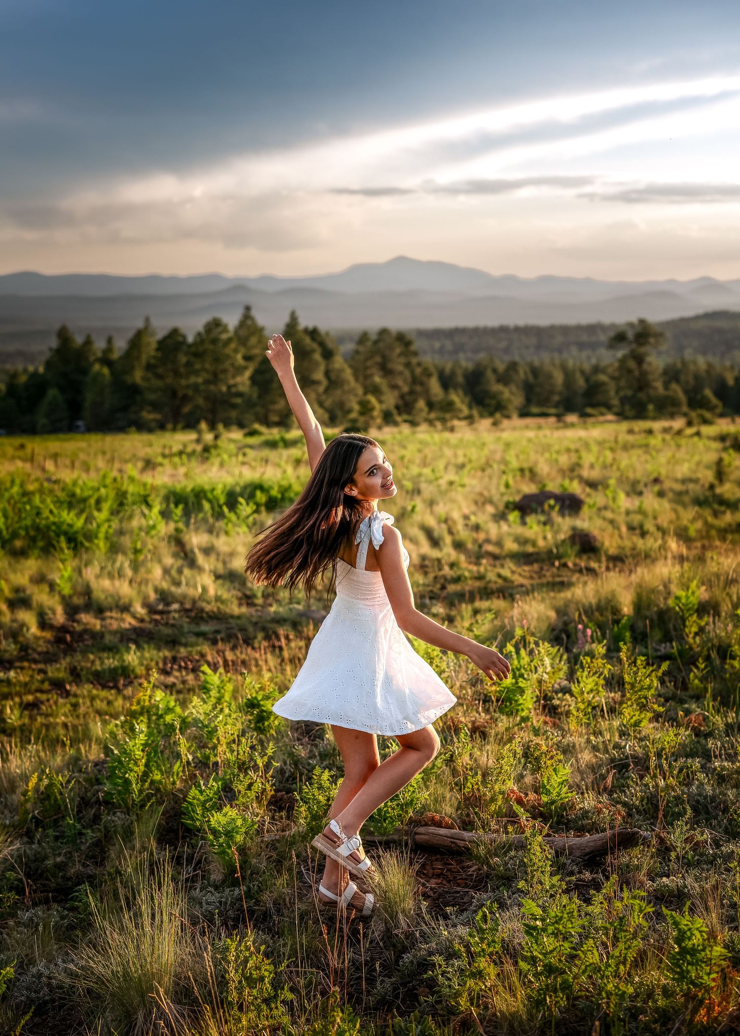 A young girl in a white dress dances joyfully in a meadow with mountains and trees in the background at sunset.