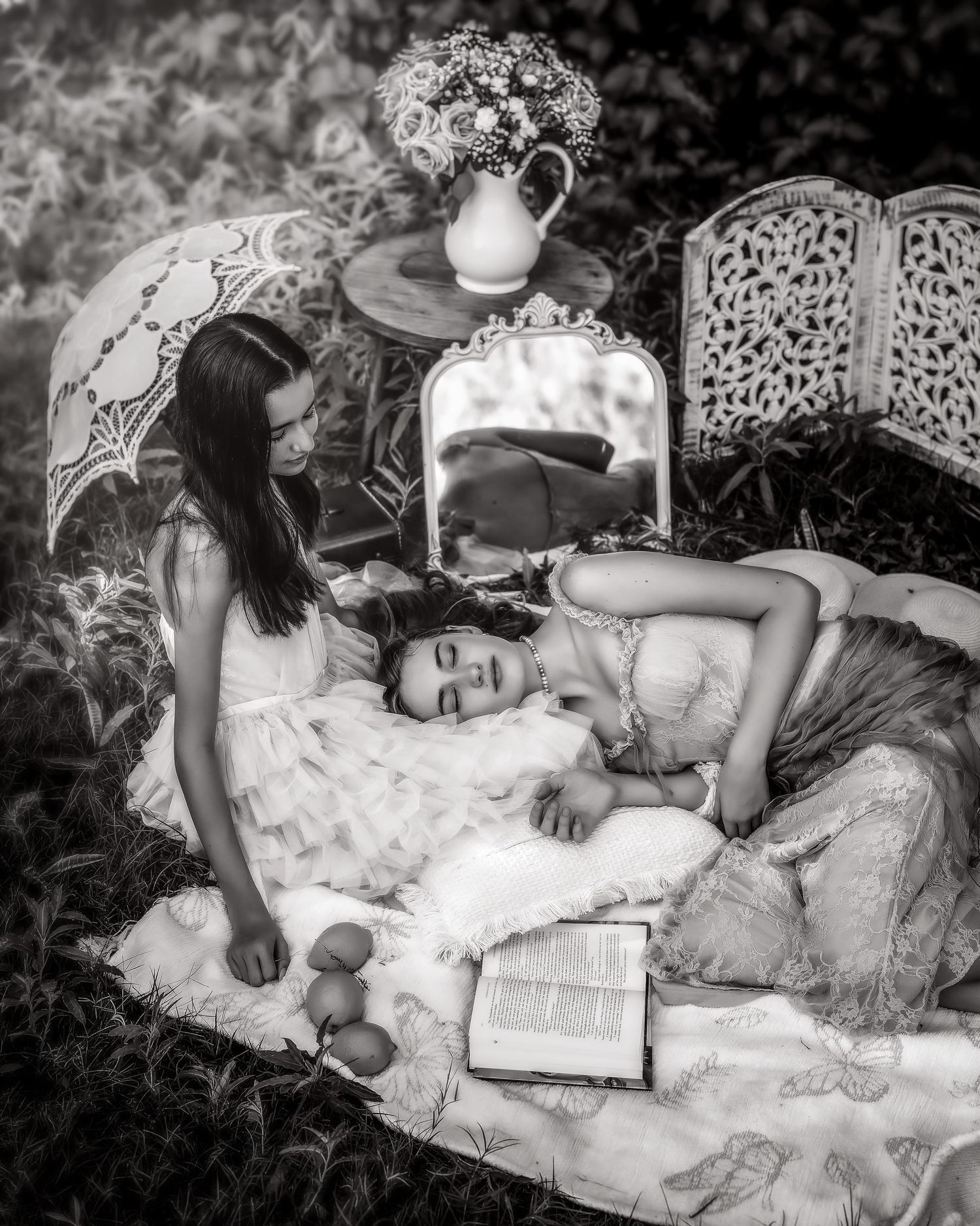 Black and white photo of two women in vintage dresses relaxing outdoors with books and flowers.
