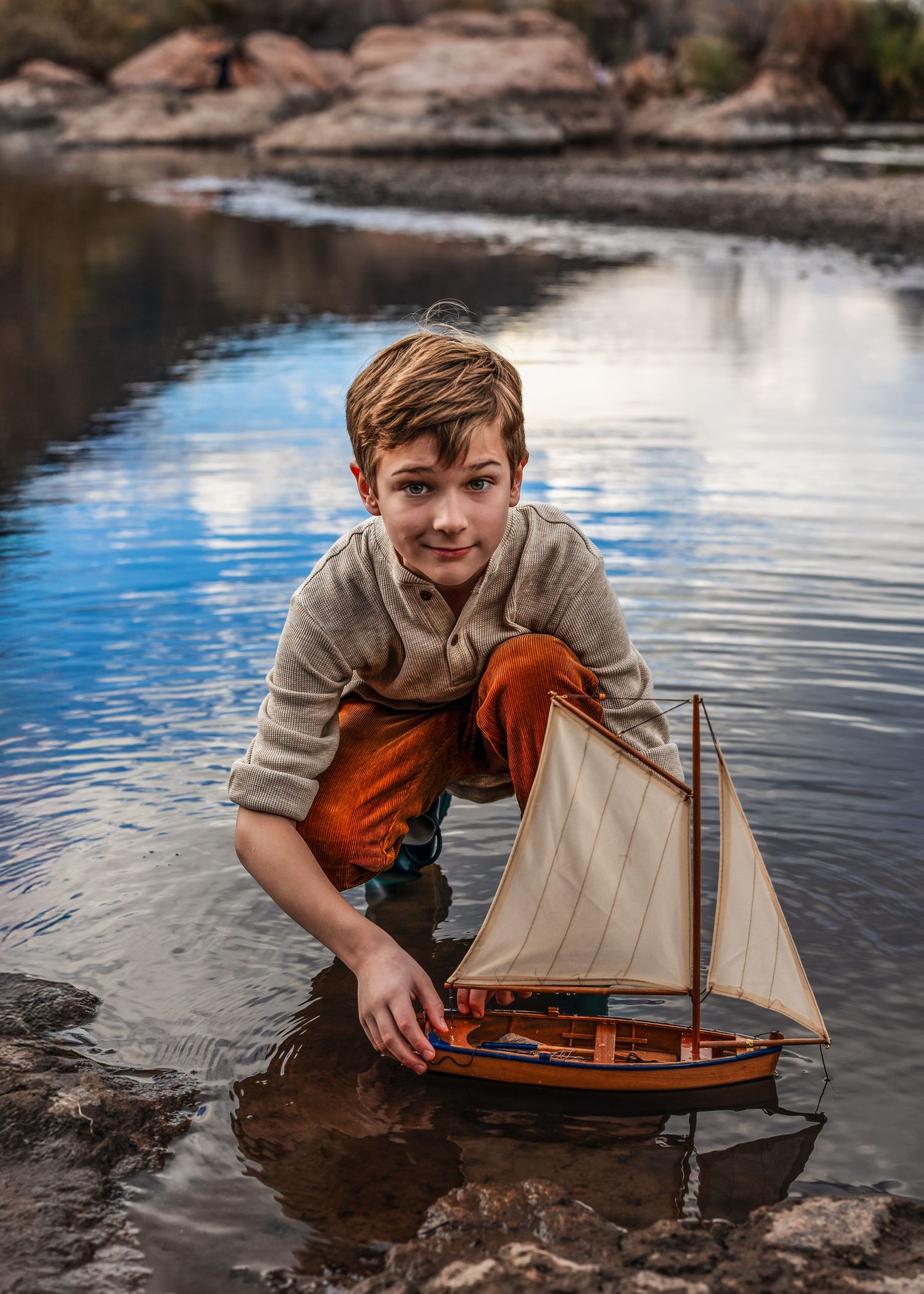 Boy crouching in shallow water playing with a wooden toy sailboat outdoors.