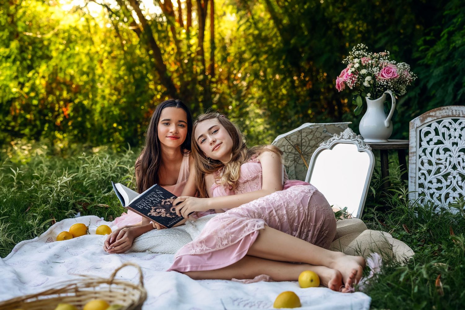 Two young women relaxing on a blanket outdoors, one reading a book, surrounded by lemons and flowers.