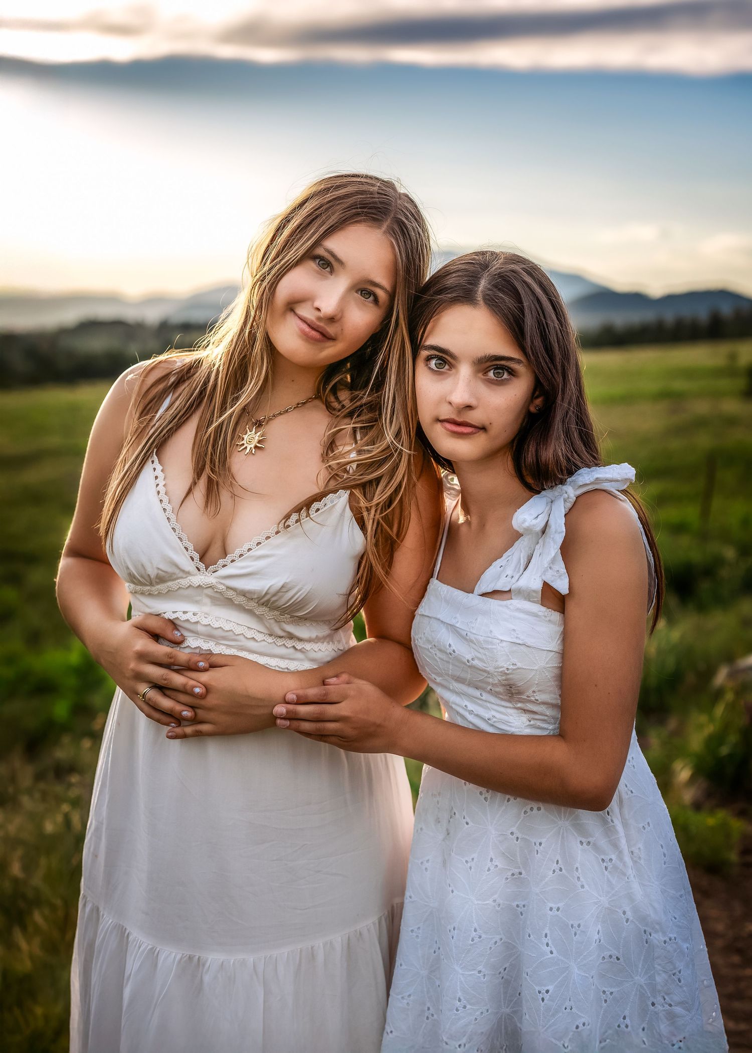 Two young women in white dresses posing together at golden hour in a scenic field.