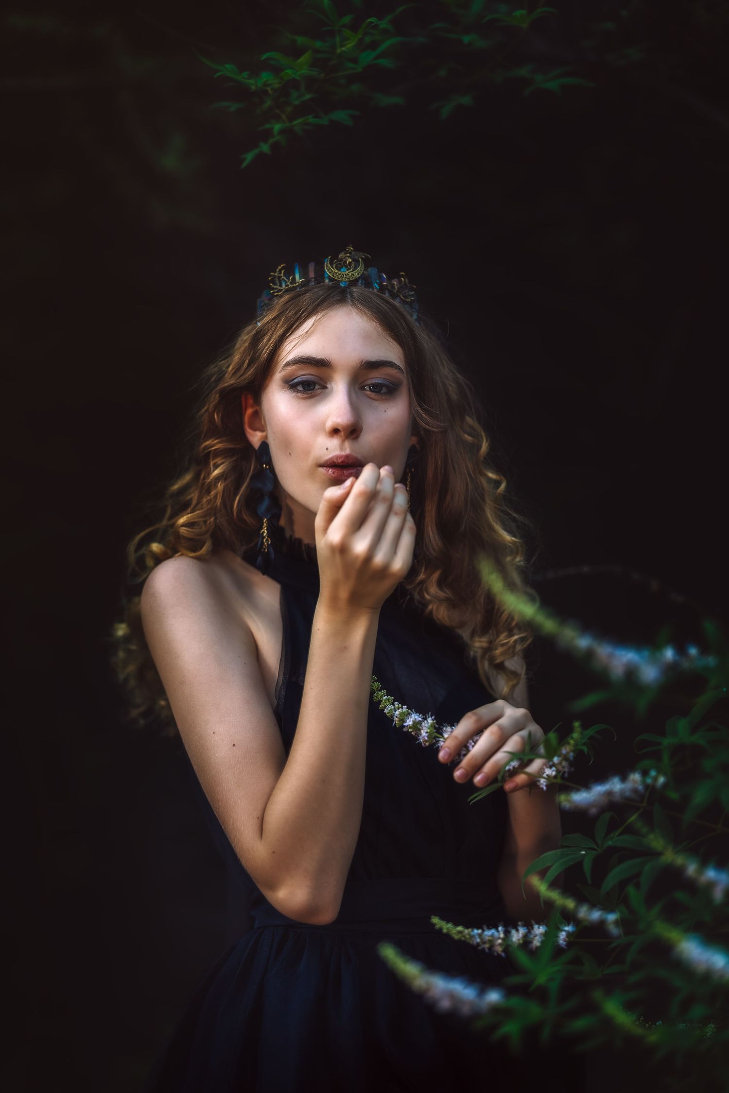 Young woman wearing a dark crown and black dress poses mystically among green forest plants in moody lighting.