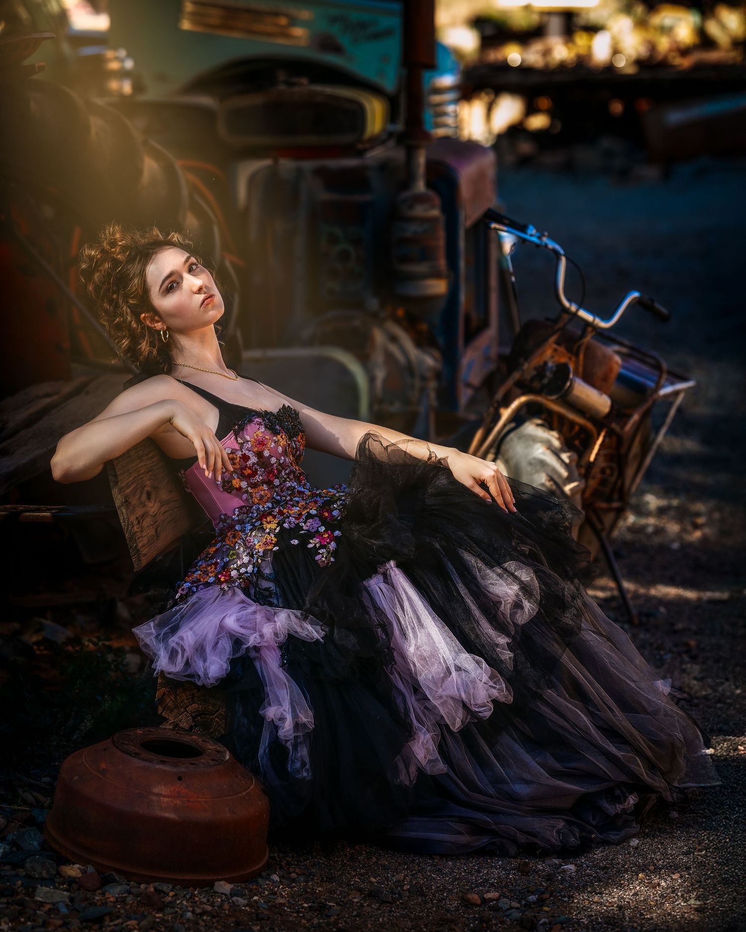 Woman in ornate black gown with floral embroidery poses dramatically among vintage rusty vehicles in golden light.