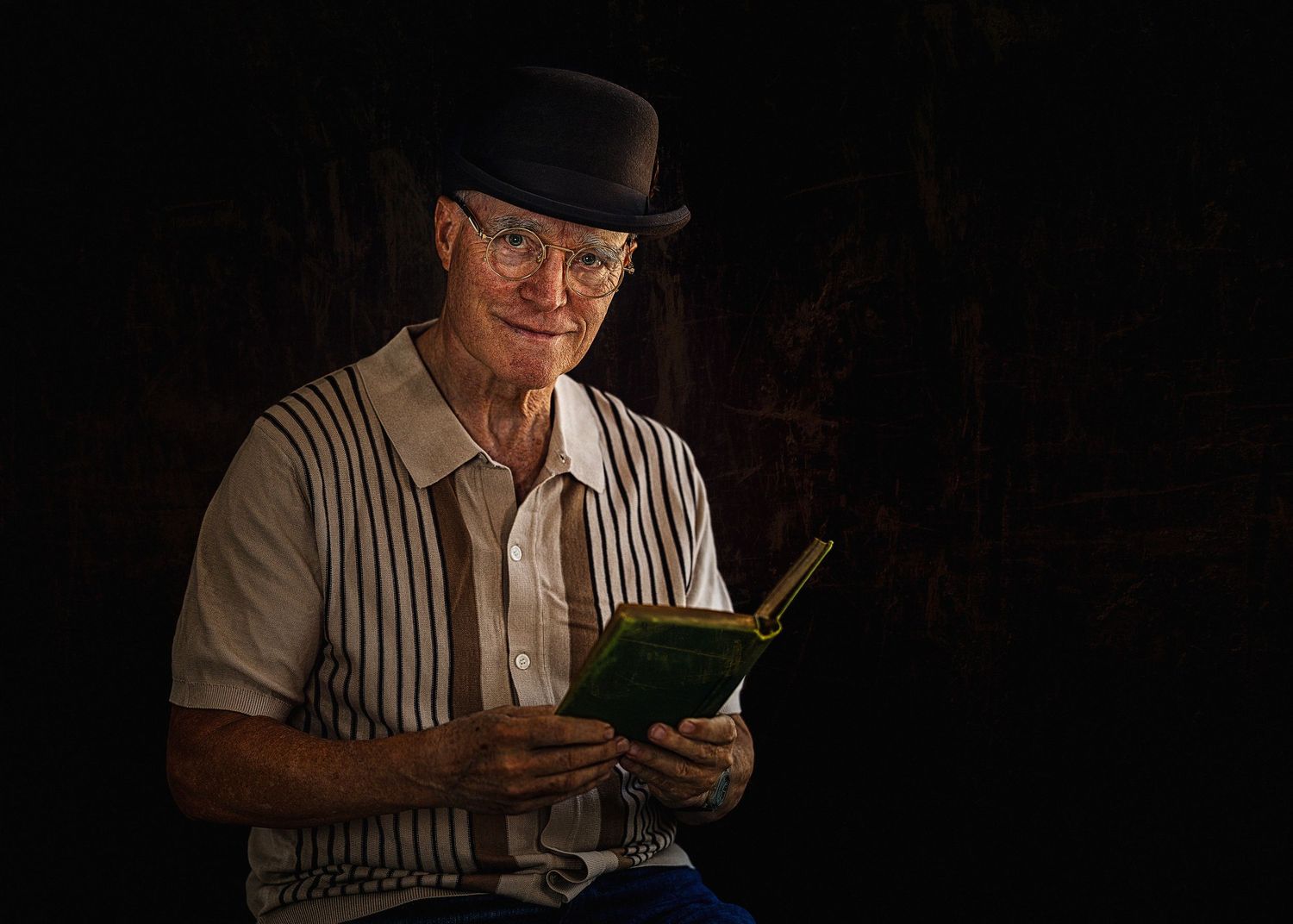 Elderly man in bowler hat and striped vest reading a green book in dim light.