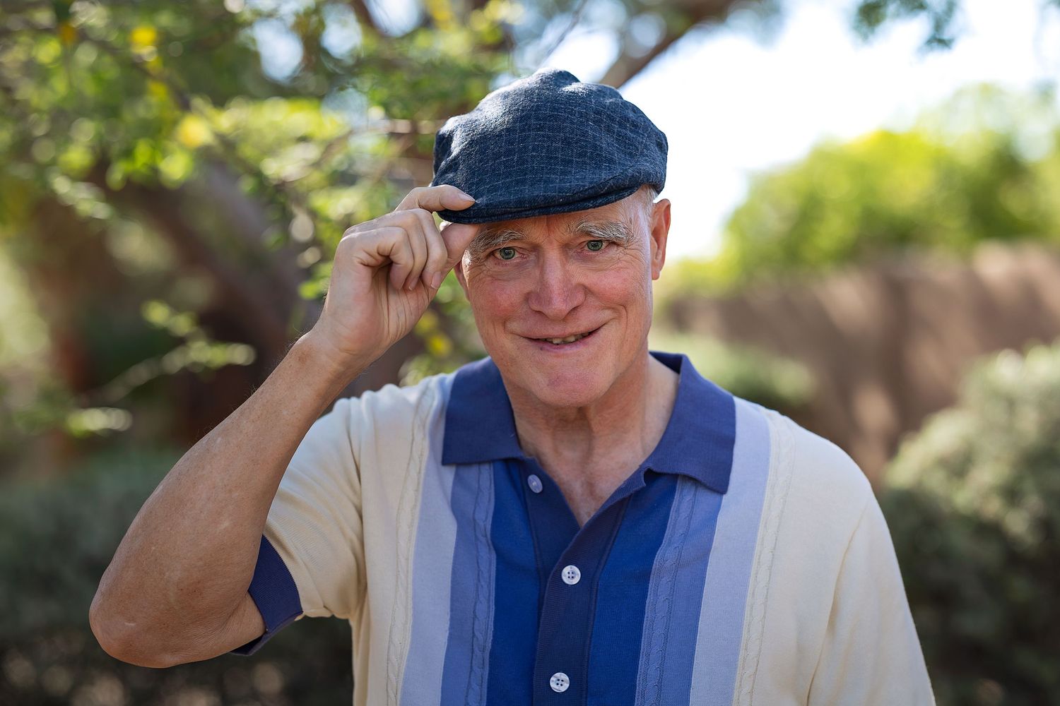 Cheerful senior man tipping his flat cap while smiling outdoors surrounded by green trees.