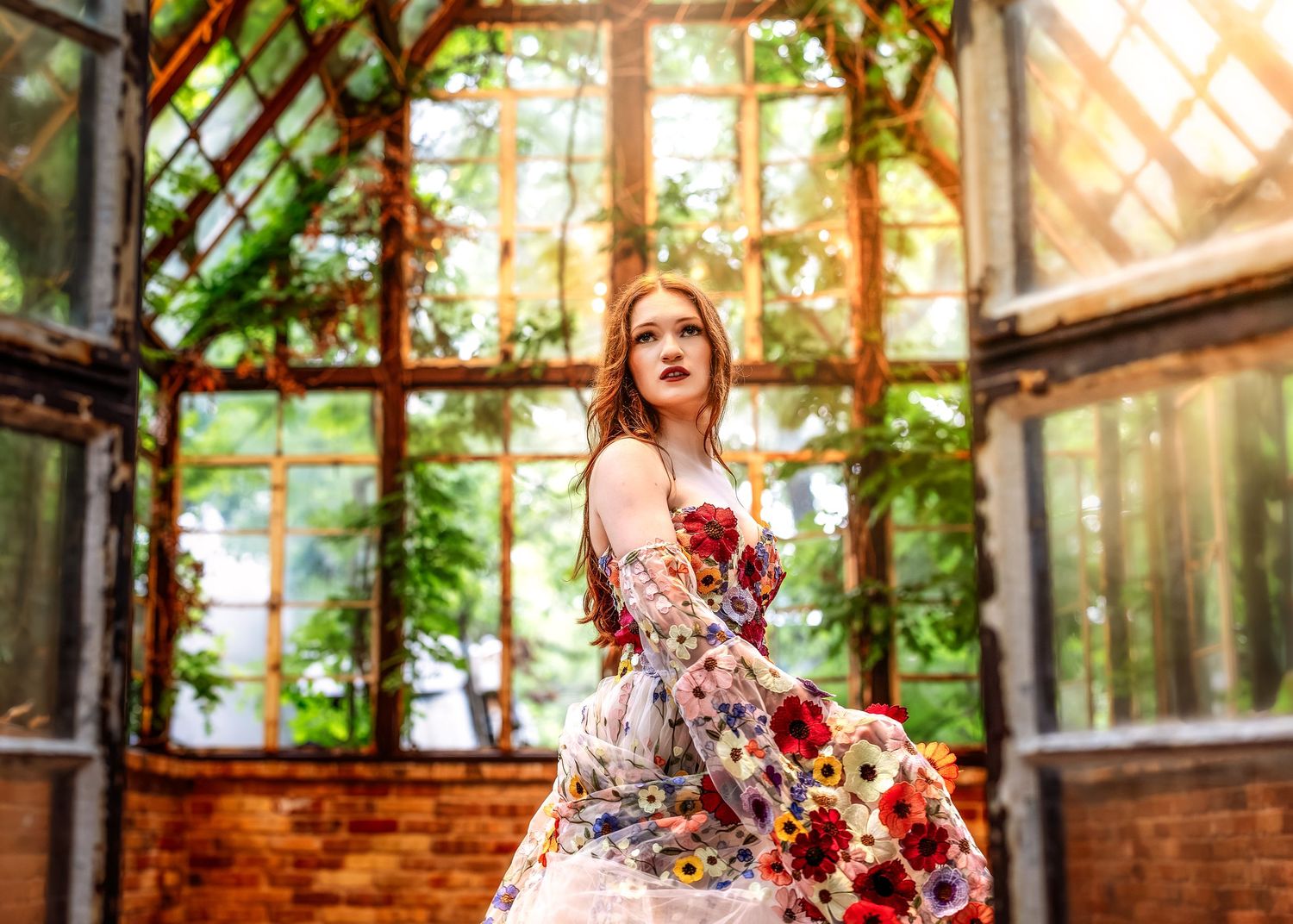 Young woman in floral dress poses gracefully in sunlit greenhouse with brick walls and lush greenery.