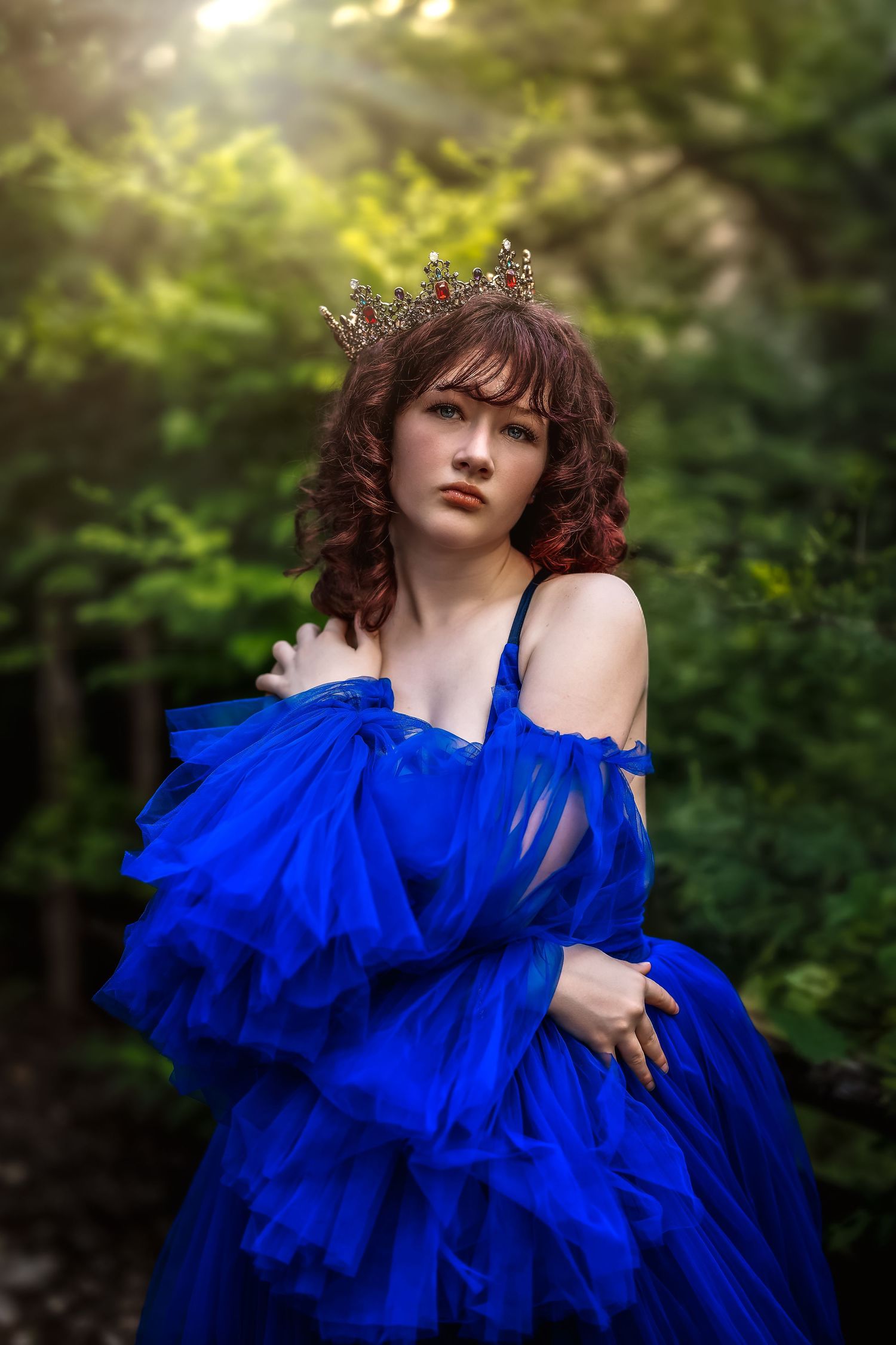 Young woman in royal blue tulle gown and gold crown poses gracefully in a sunlit forest setting.