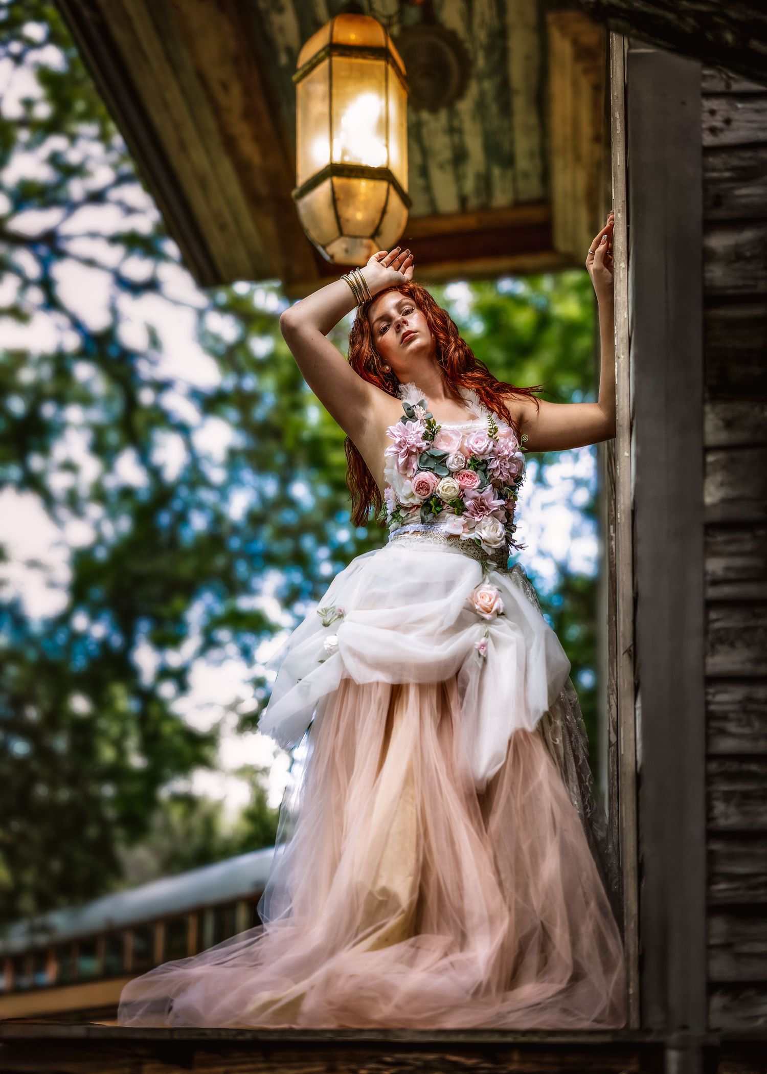 A woman in a floral tulle gown poses gracefully under a vintage lantern on a rustic wooden porch.