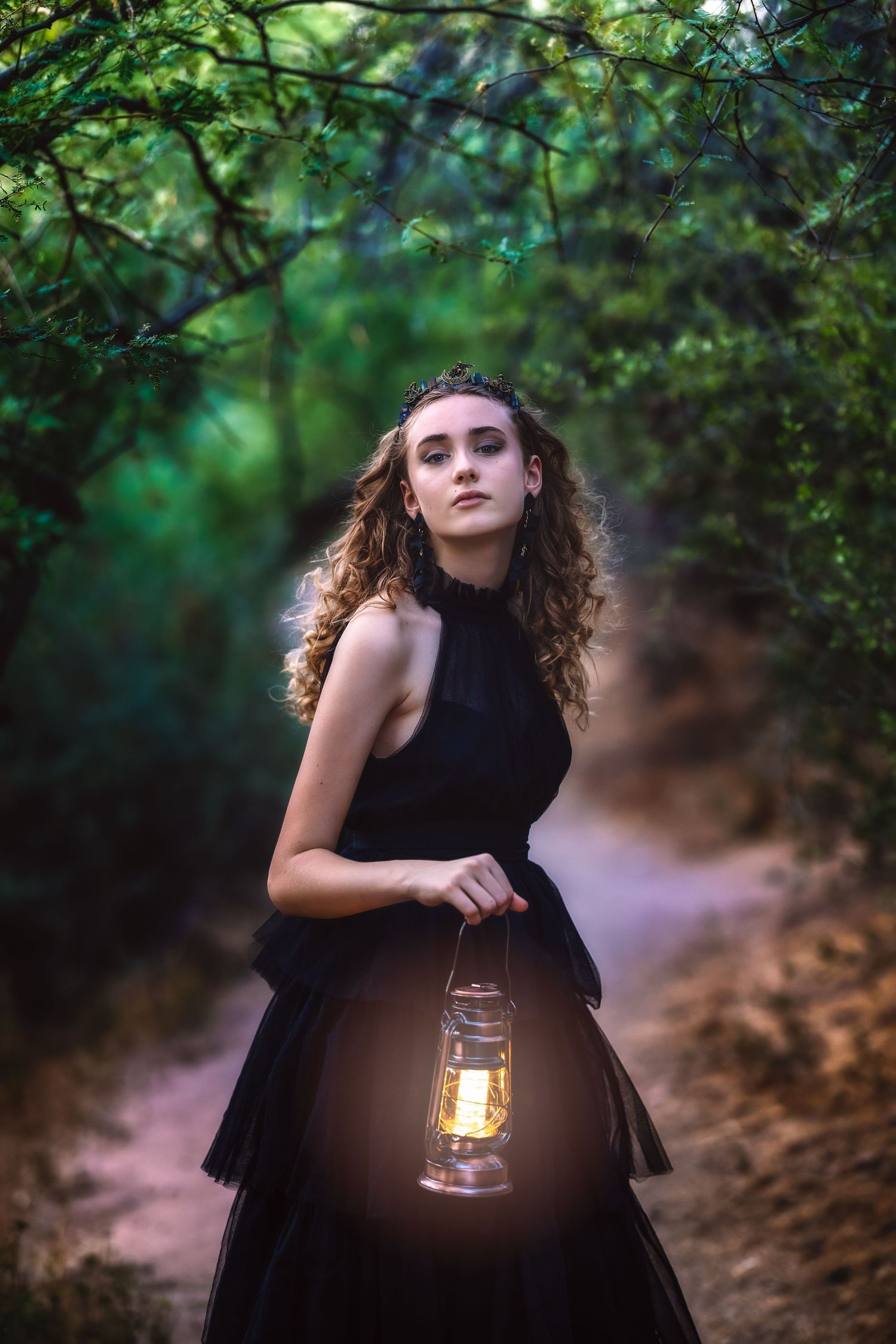 Young woman in black dress holds glowing lantern on forest path at dusk.