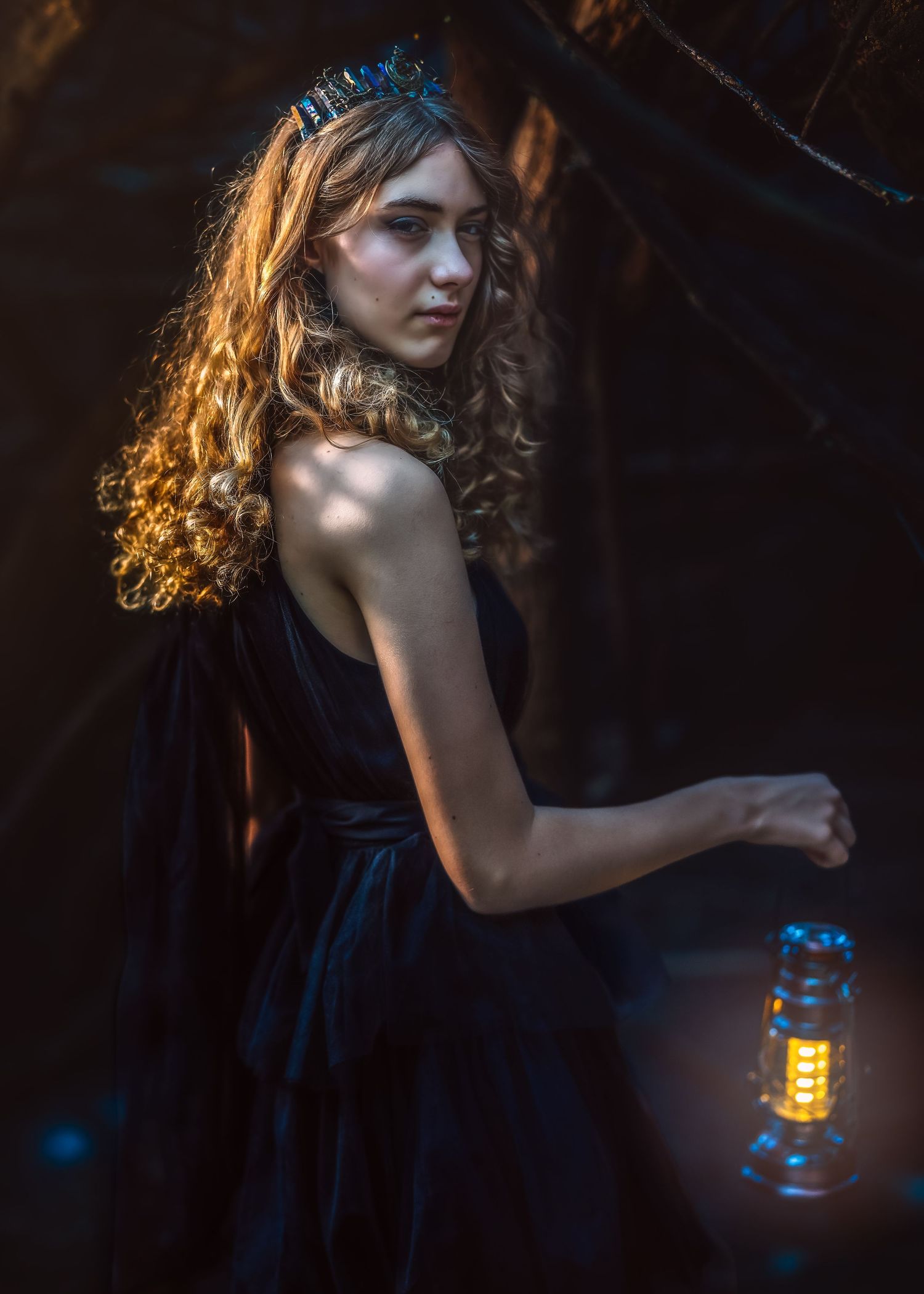 Young woman in black dress holding a glowing lantern in a dark forest, wearing a decorative crown.