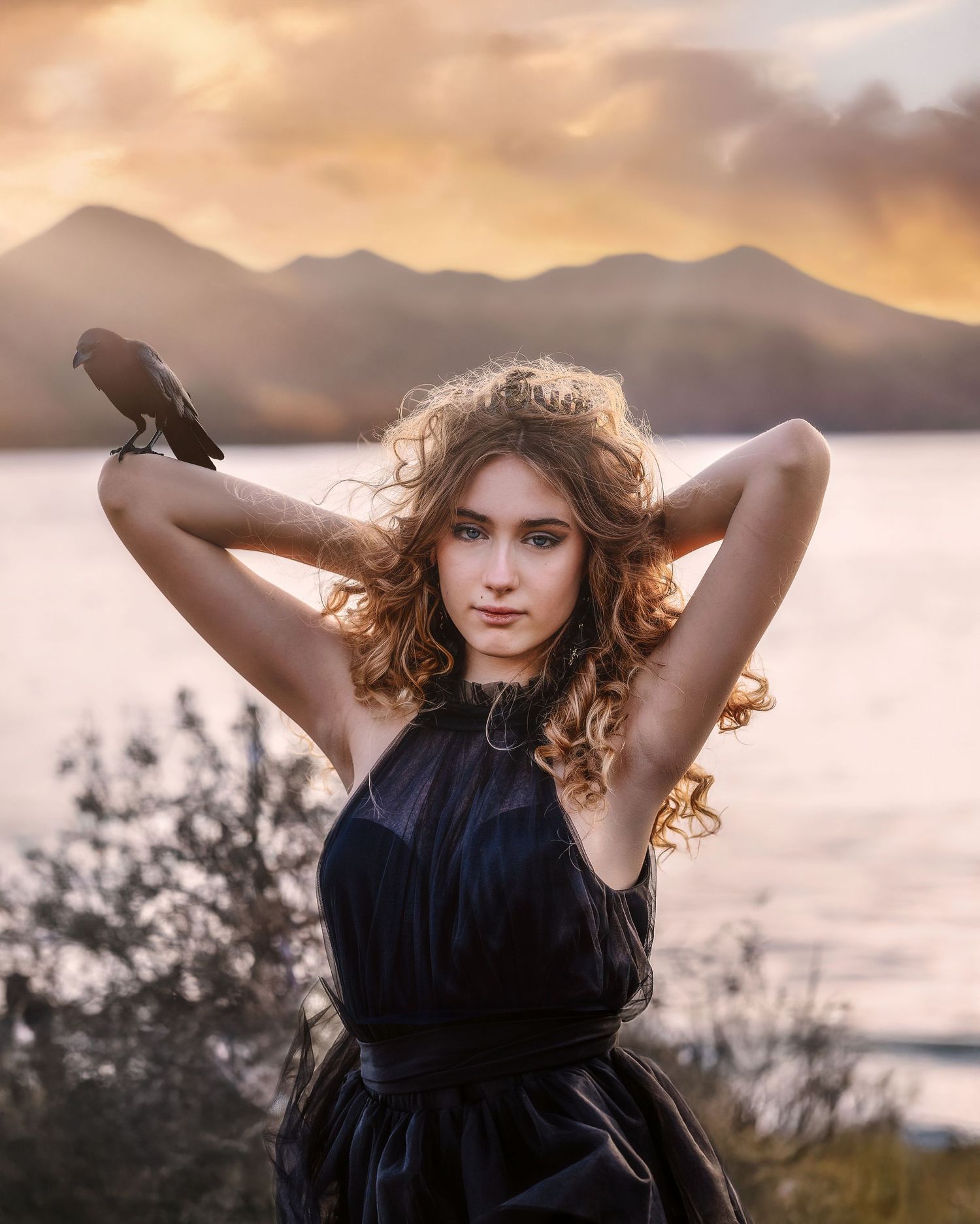 Young woman in black dress with a bird on her wrist poses against a golden sunset over mountains and lake.