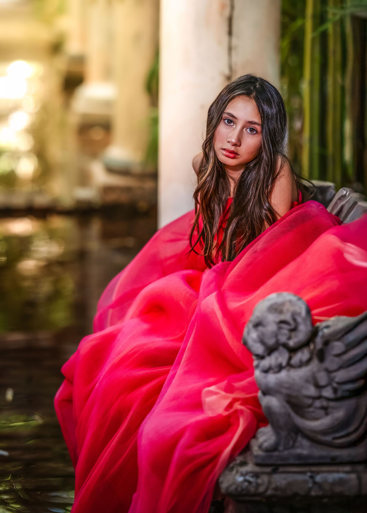 Young woman in flowing red gown seated by garden pond with cherub statue.
