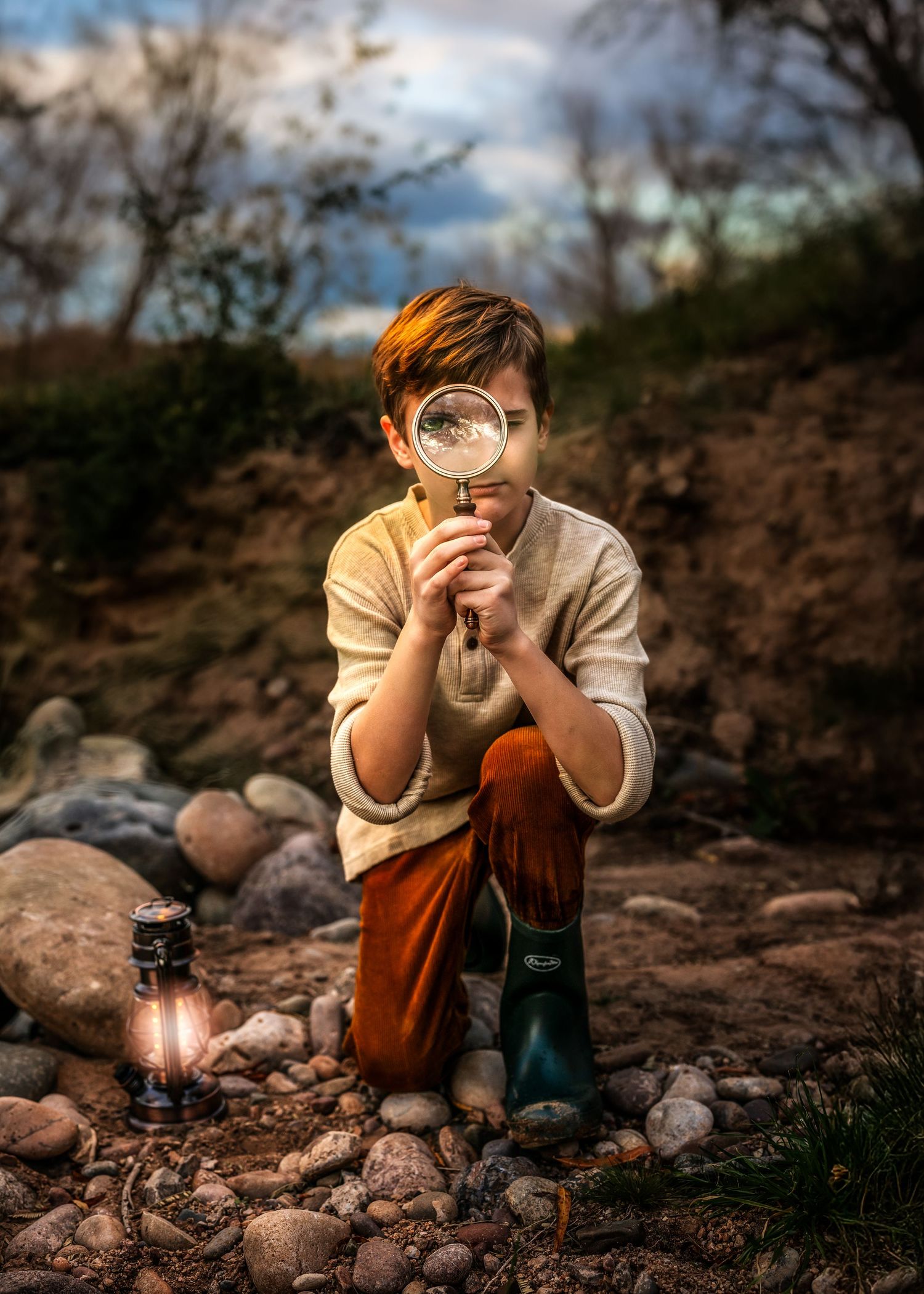 Boy kneeling outdoors holding magnifying glass with lantern beside him on rocky ground at dusk.