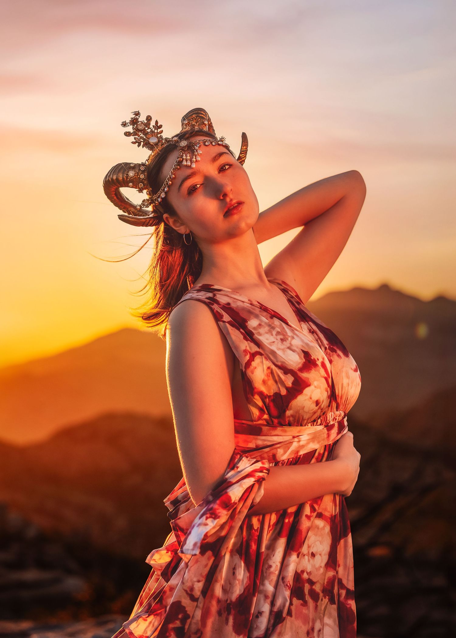 Woman in floral dress with ram horn crown poses at golden sunset in mountains.