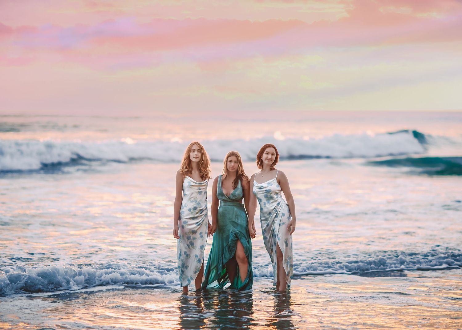 Three women in flowing dresses standing in ocean waves at sunset.