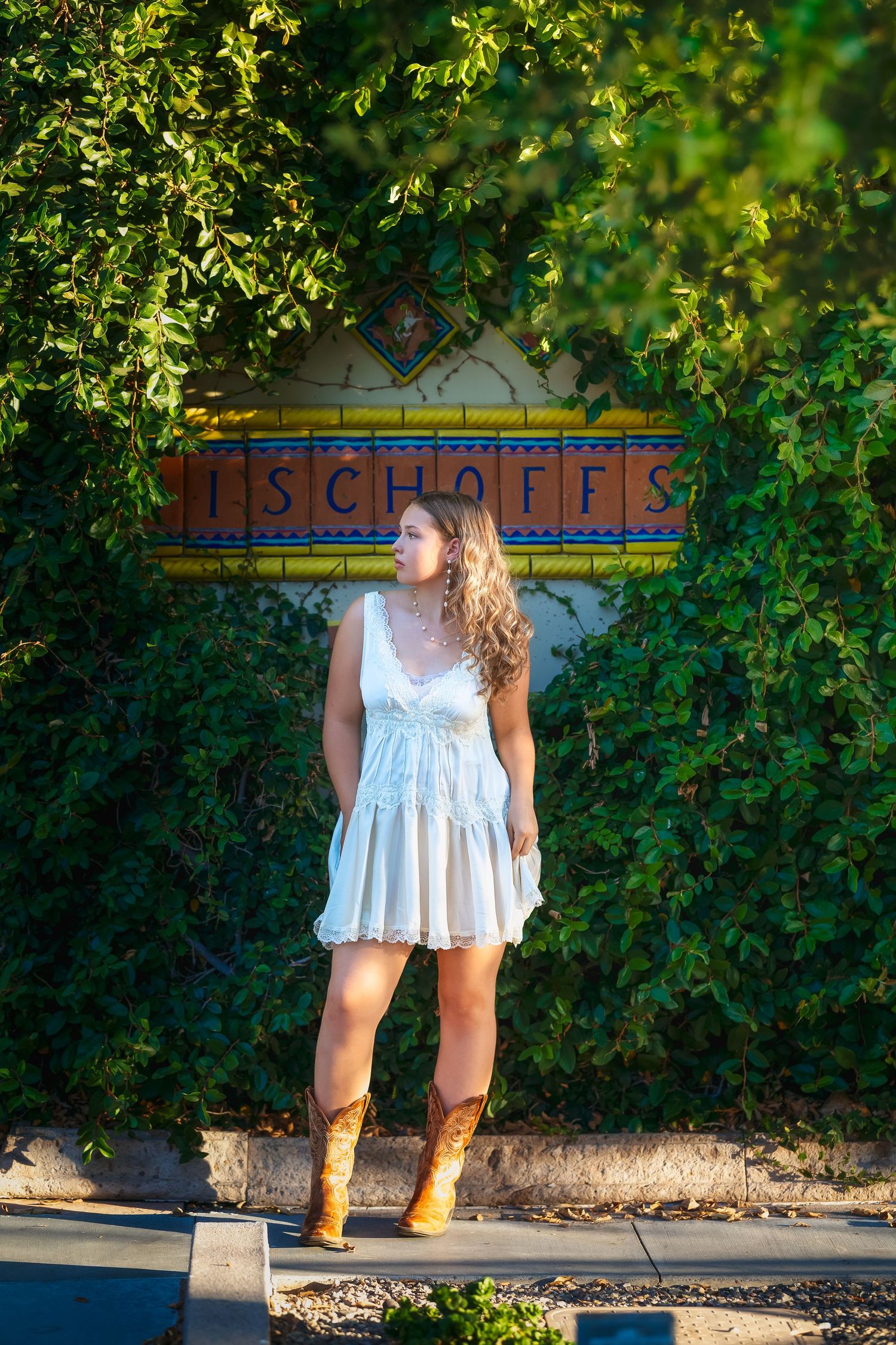 Young woman in white dress and cowboy boots stands before a colorful tiled sign surrounded by lush green ivy.