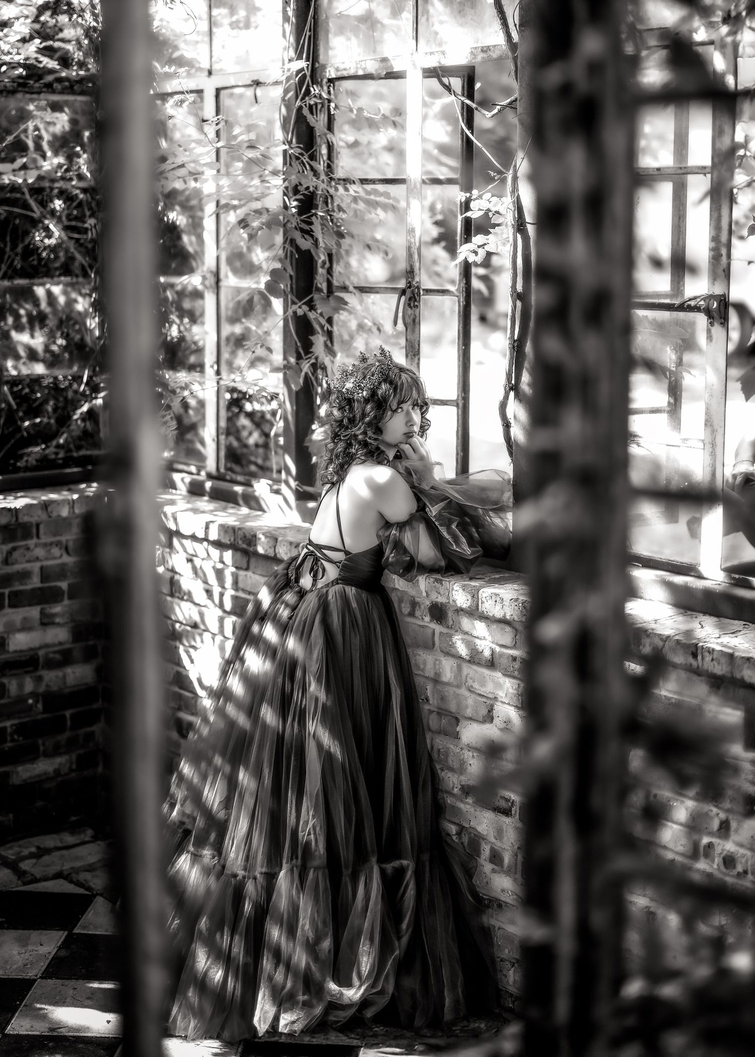 Black and white photo of a woman in an elegant ball gown standing by a window in a rustic brick greenhouse.
