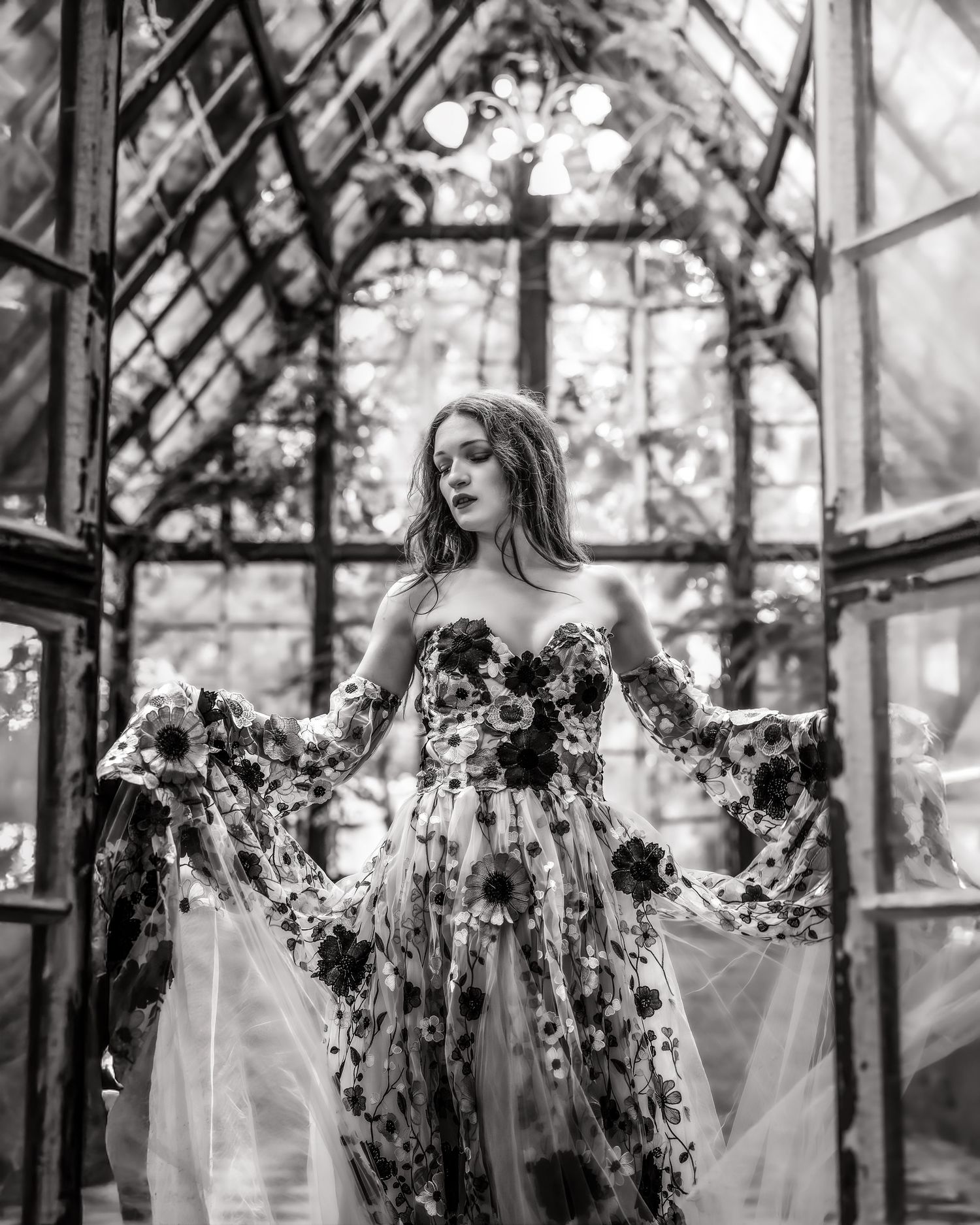Woman in floral strapless gown poses in black and white photo inside vintage greenhouse with arched glass ceiling.