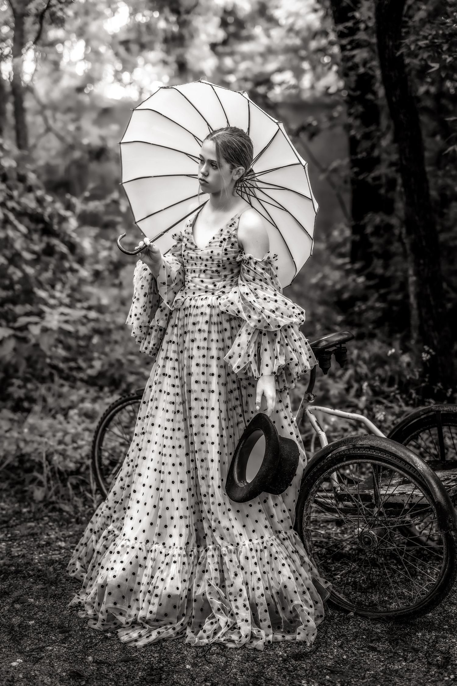 Woman in polka dot gown holding parasol and hat beside vintage bicycle in forest, black and white photo.