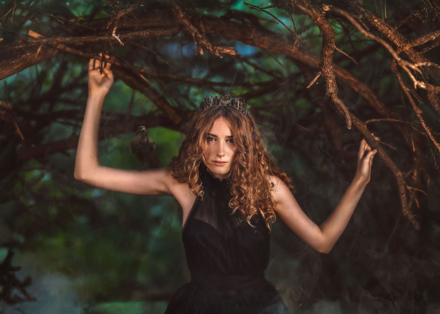A young woman with curly hair in a black dress poses among dark tree branches in a moody forest setting.