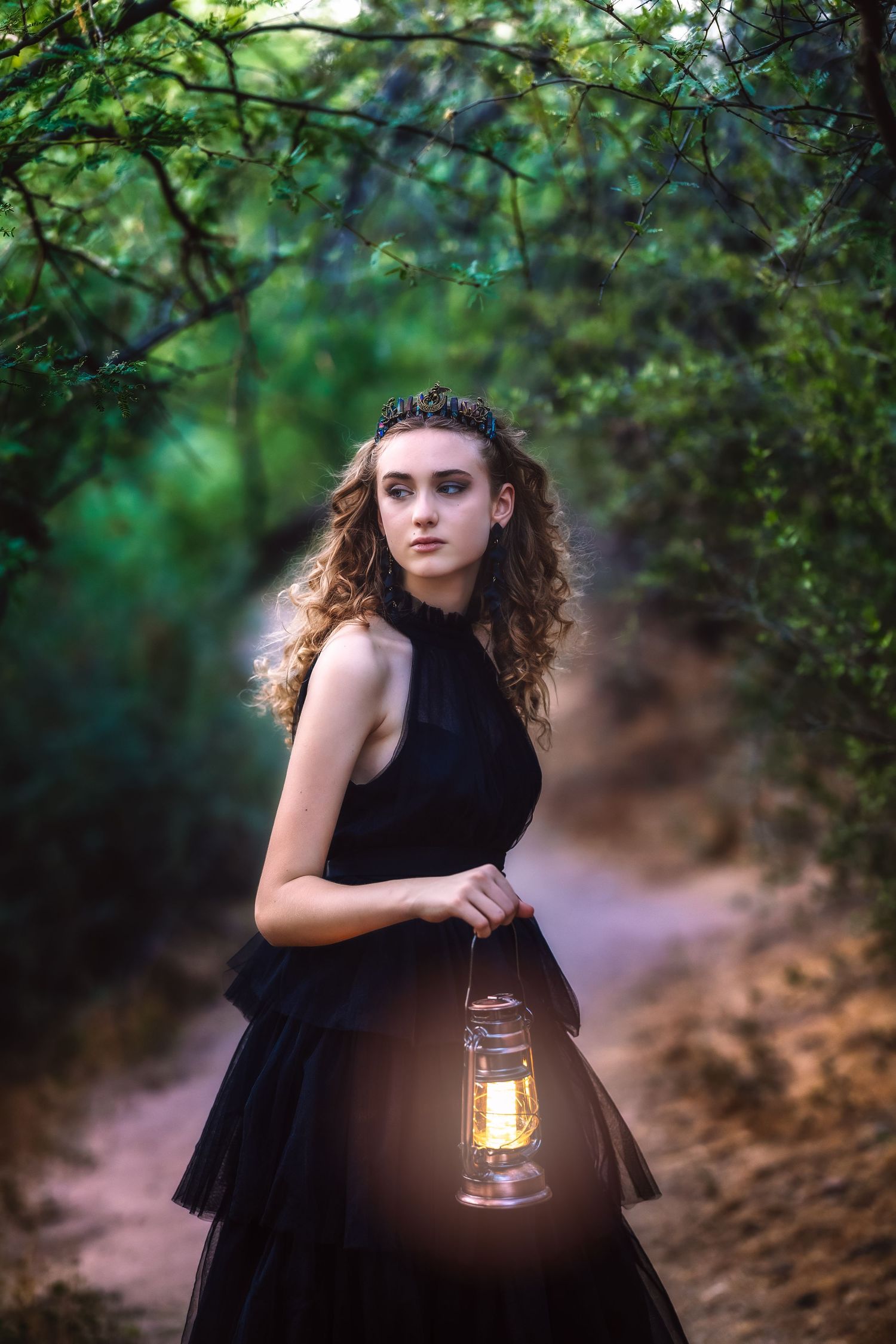 Young woman in black dress holding glowing lantern on forest path at dusk.