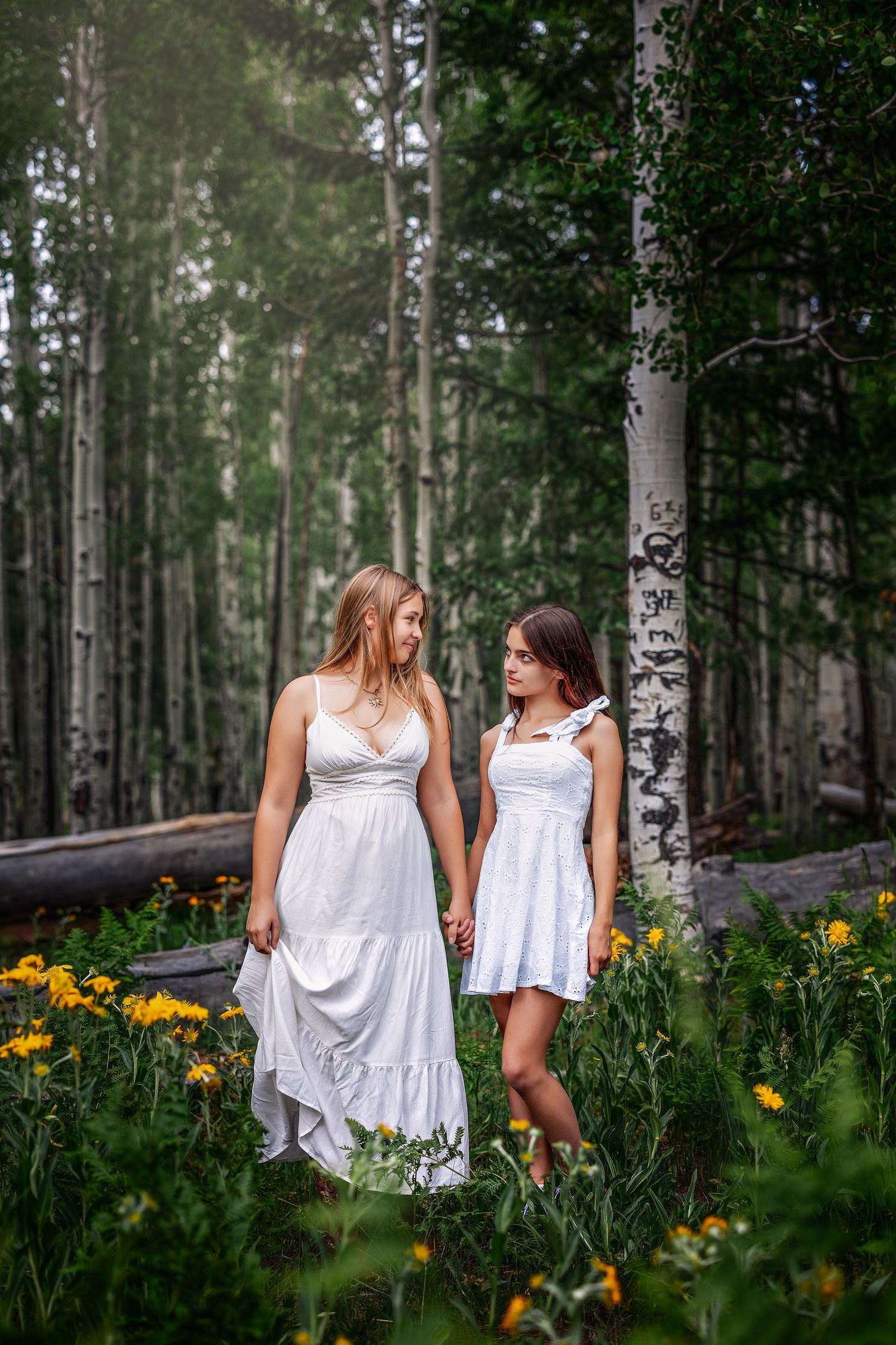 Two young women in white dresses hold hands among wildflowers in a birch forest.