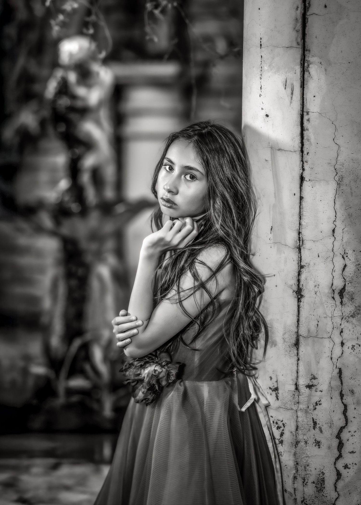 Black and white portrait of girl in elegant gown leaning against ornate column with statue.