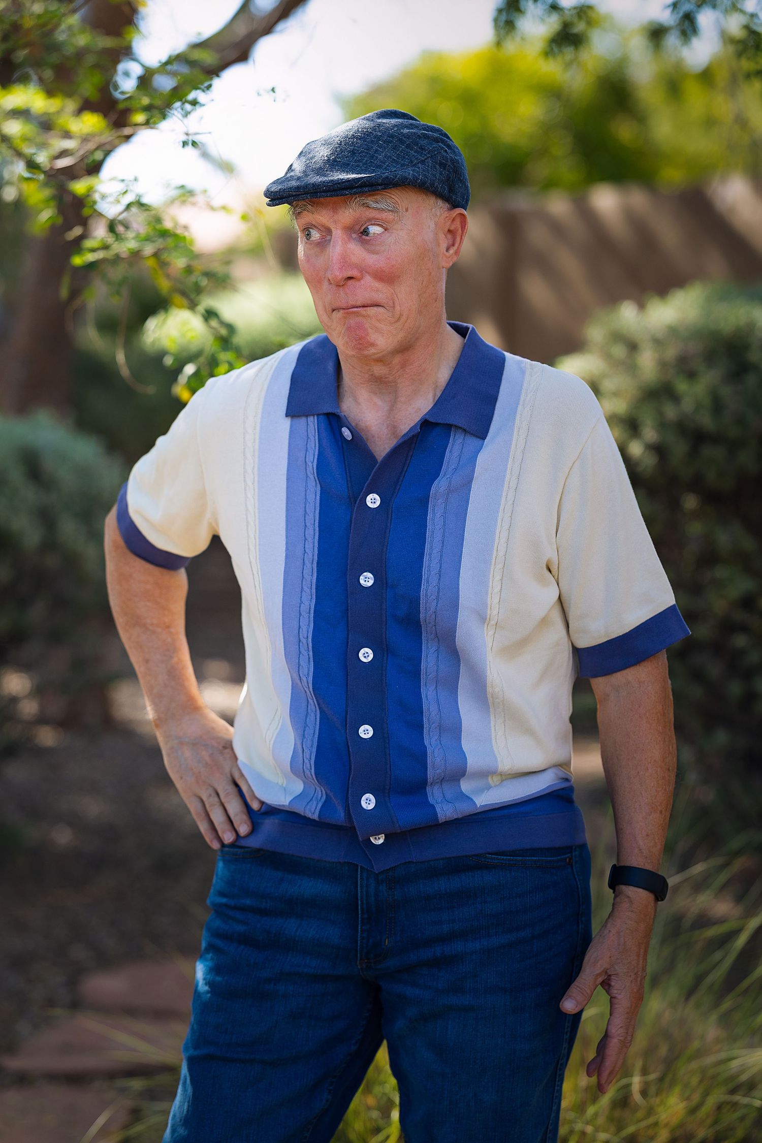Senior man in a blue striped polo shirt and flat cap making a funny face outdoors in a garden.