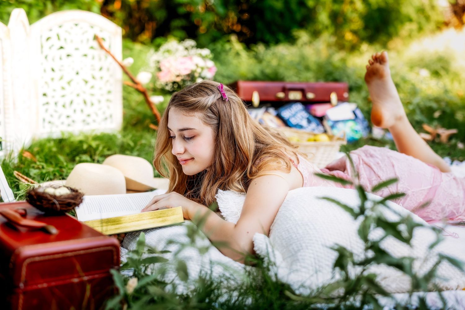 Young girl in white dress lying on a blanket outdoors reading a book in a garden setting.