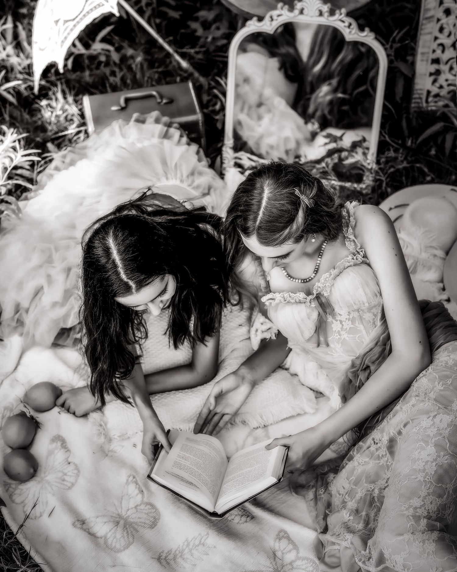 Black and white photo of two girls in fancy dresses reading a book together outdoors.