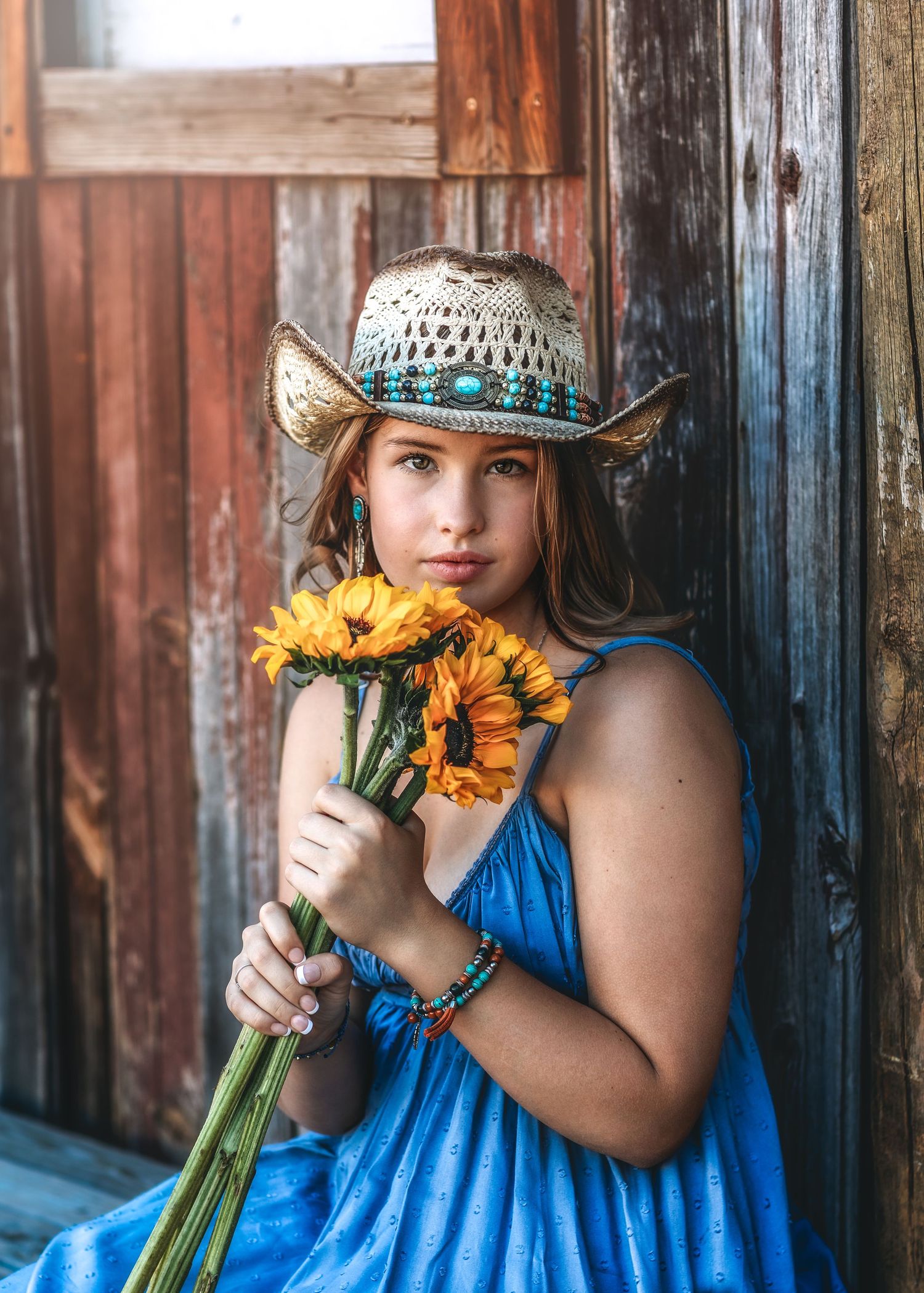 Young woman in blue dress and turquoise cowboy hat holding orange sunflowers outdoors.