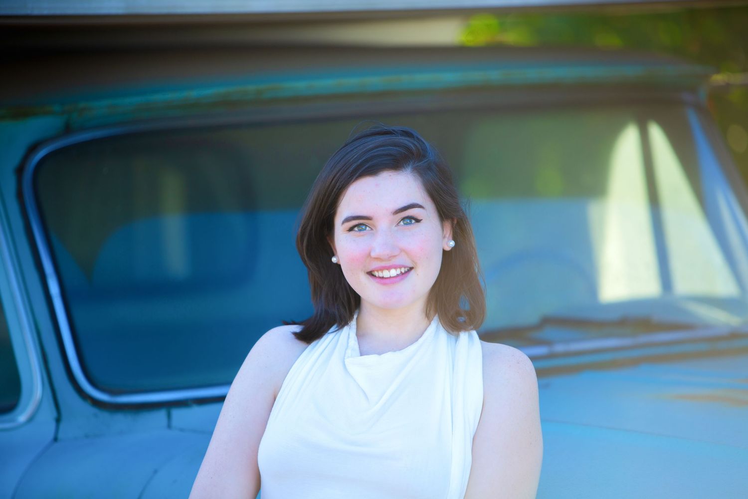 Smiling young woman in white dress leaning against classic blue vintage truck outdoors.