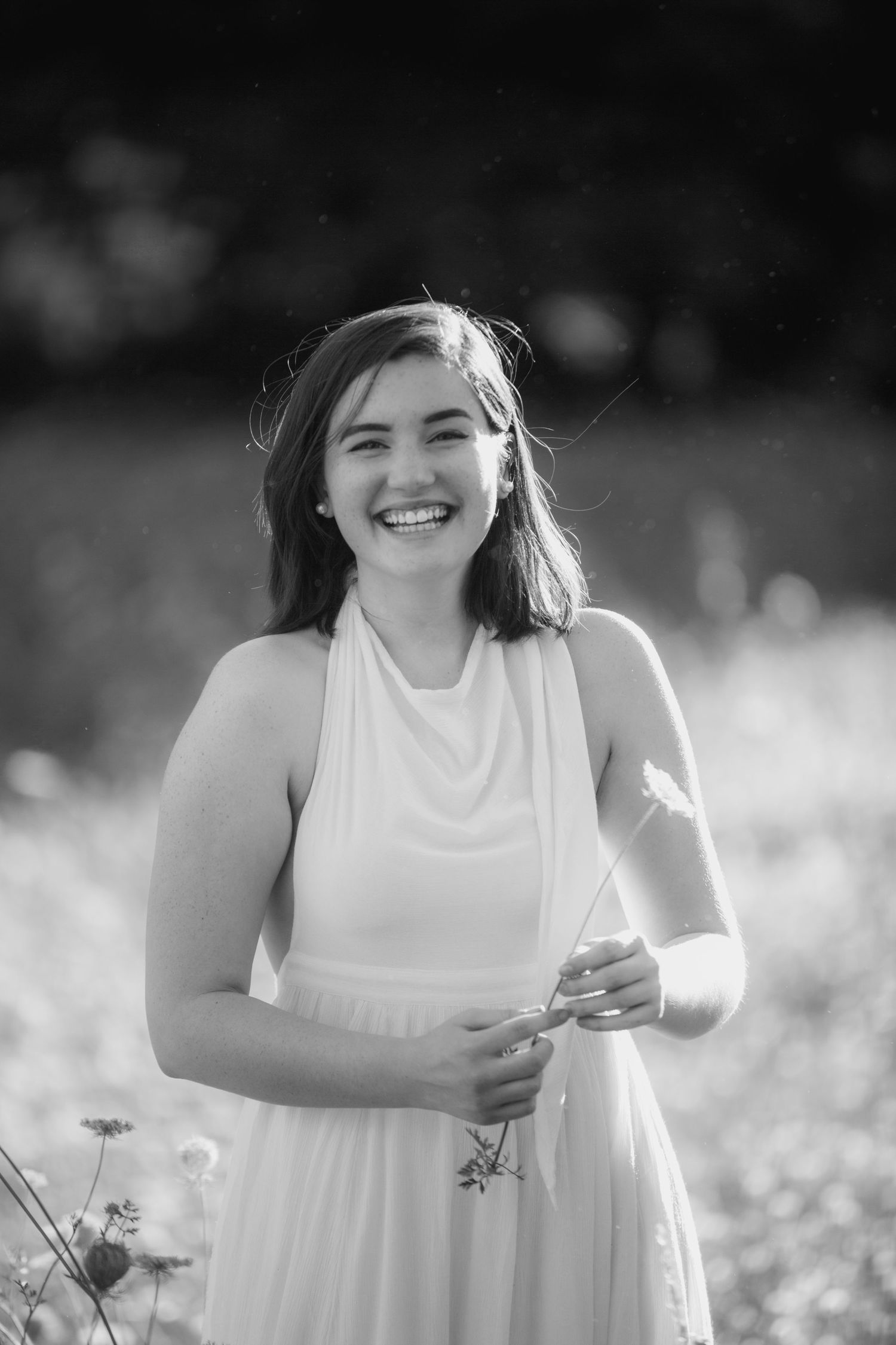 Black and white portrait of a smiling young woman in a white halter dress holding a wildflower in a sunny field.