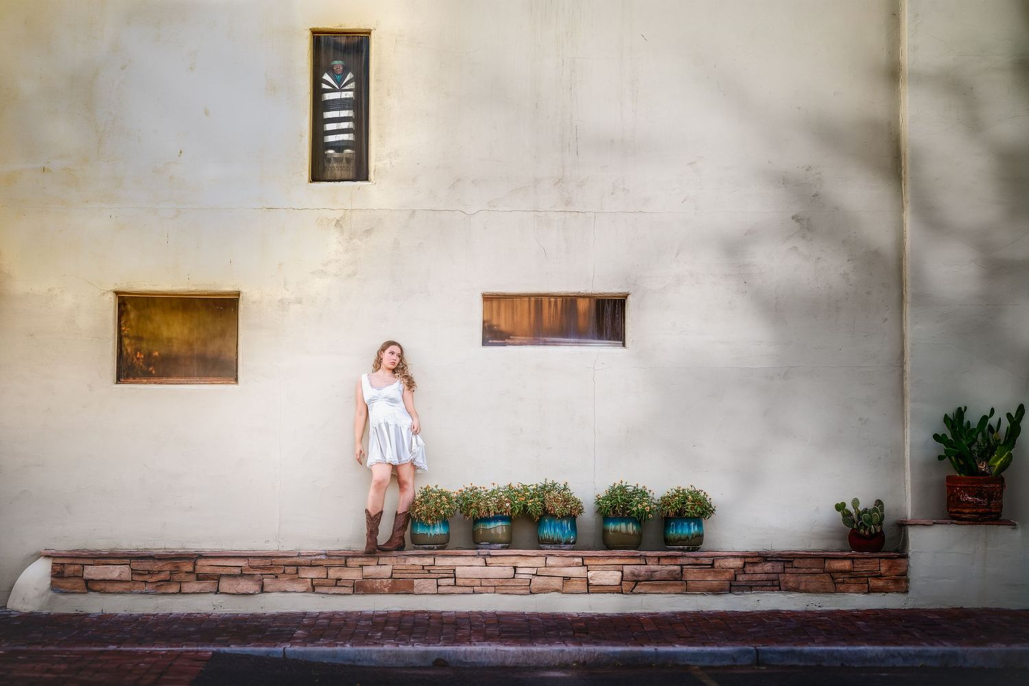 Woman in white dress standing beside a rustic brick planter with flowers against a textured beige wall.
