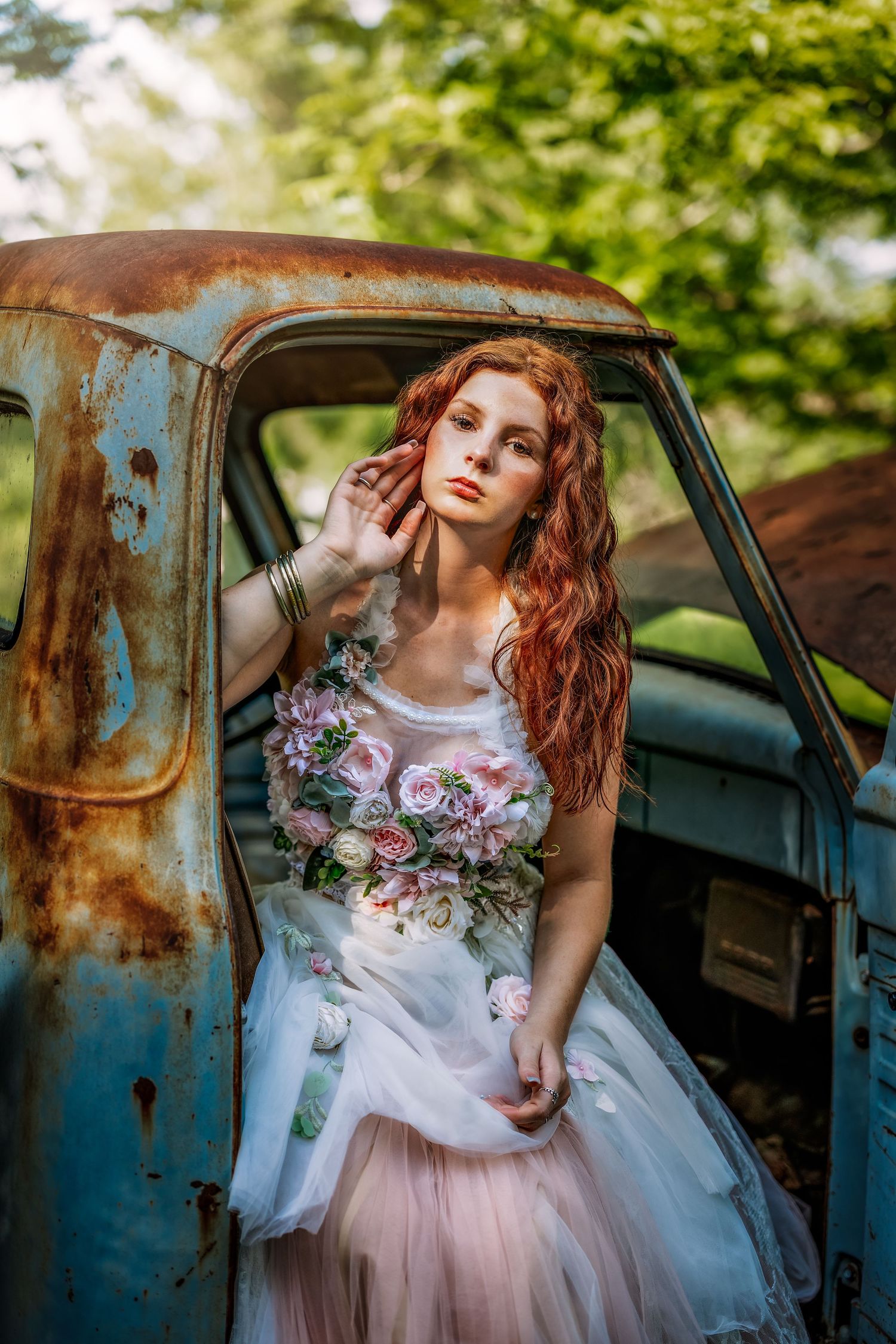 A red-haired woman in a floral wedding gown poses gracefully beside a rusty vintage truck outdoors.