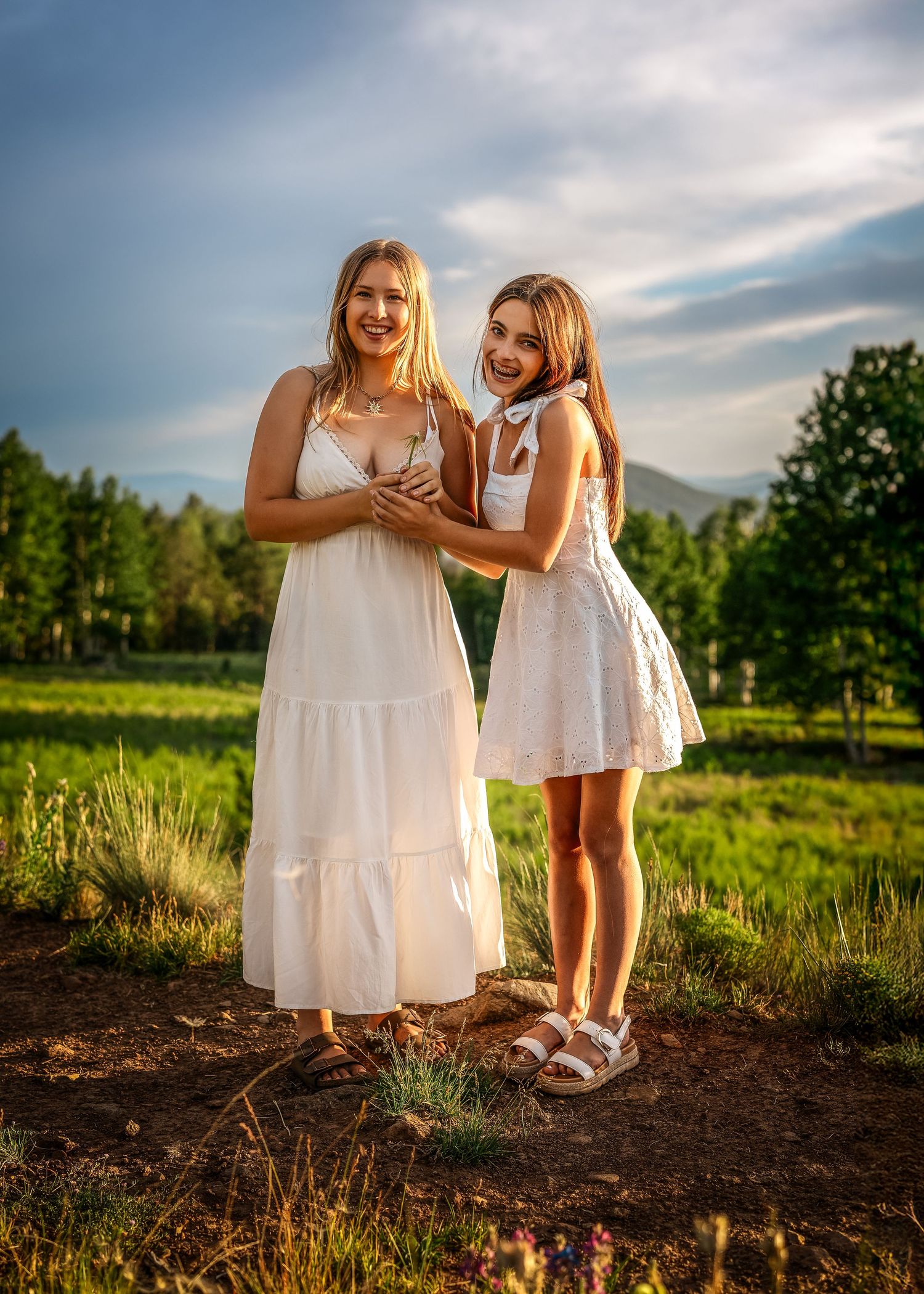 Two smiling girls in white dresses pose outdoors in a sunny green field with trees and mountains behind them.
