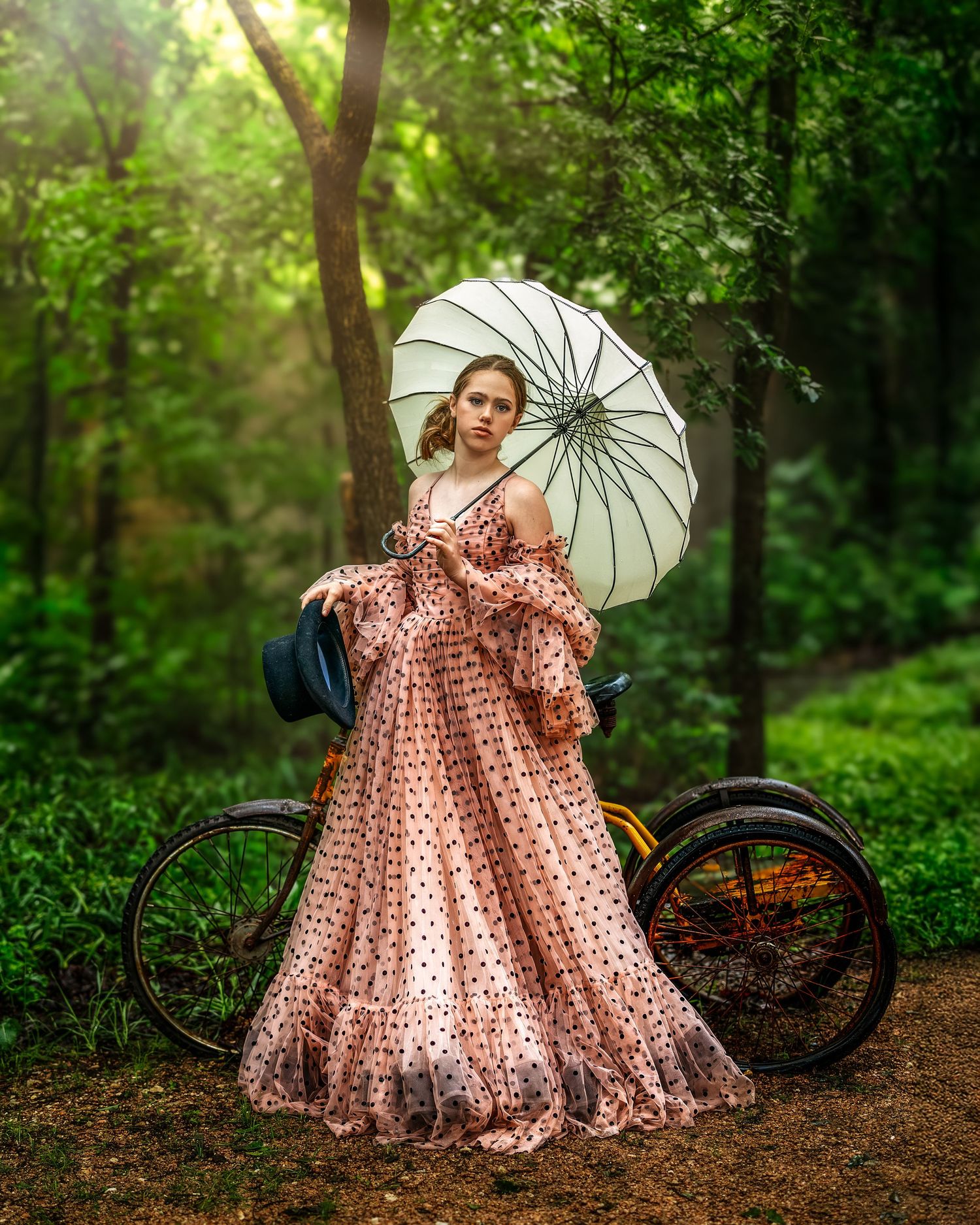 Young woman in pink polka dot gown holding white parasol beside vintage bicycle in lush green forest.