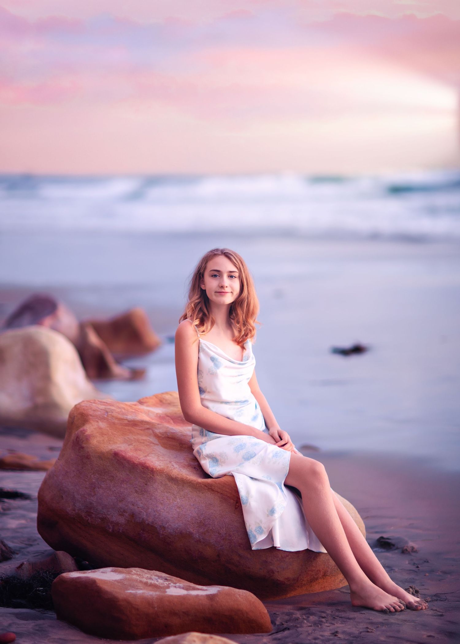 Young woman in white dress sits on sandy beach rock under soft pink sunset sky.