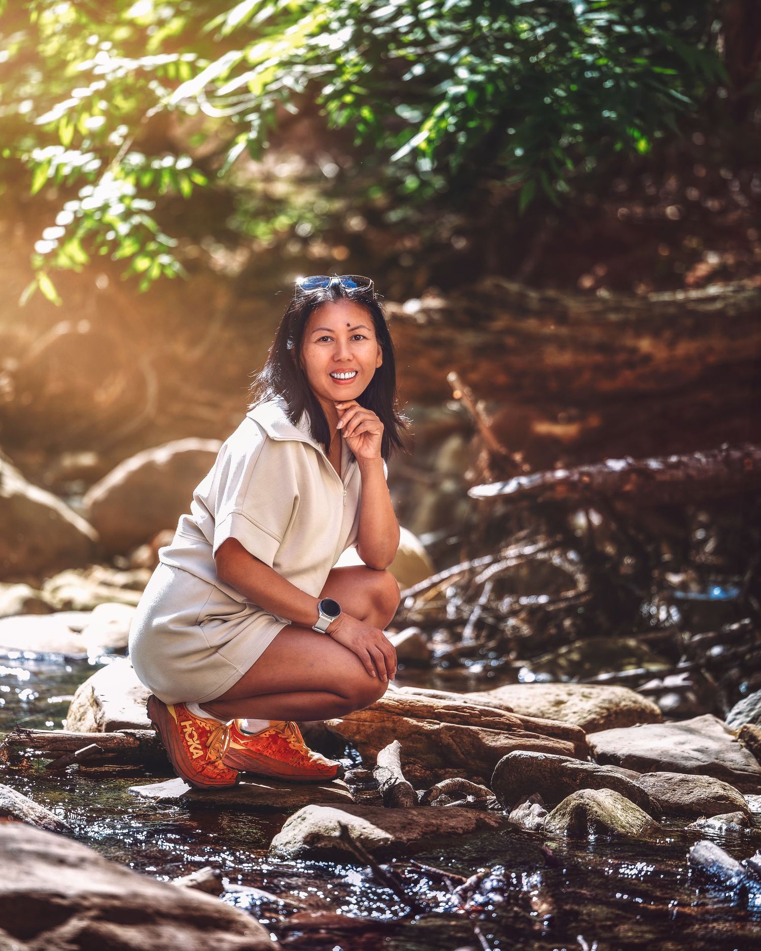 Smiling woman crouching on rocks by a stream in a lush green forest, wearing a beige outfit and orange shoes.