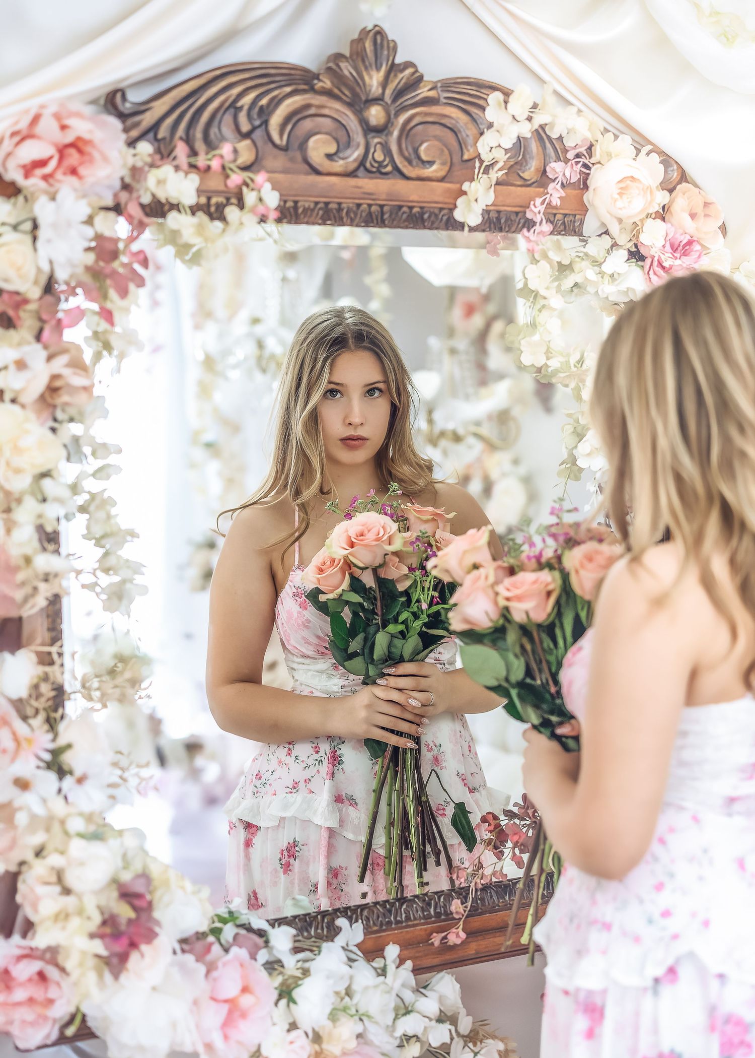 Young woman in floral dress holding pink roses bouquet reflected in ornate flower-adorned mirror.