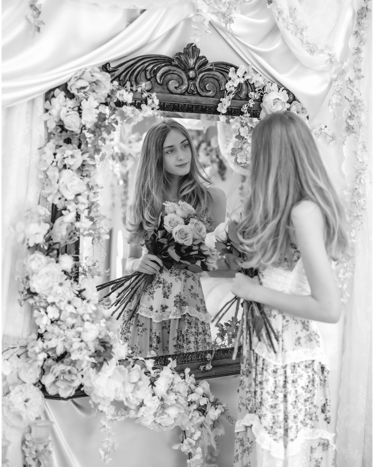 Young woman in floral dress holding bouquet reflected in ornate flower-adorned mirror, black and white photo.