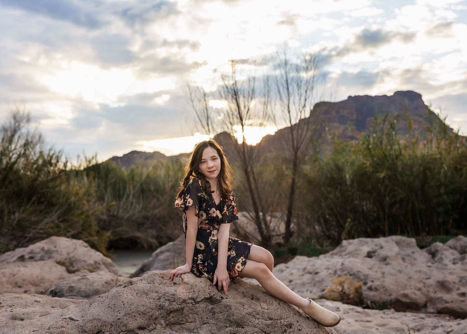 Teen girl in floral dress sitting on rocks with mountain and sunset sky behind her.