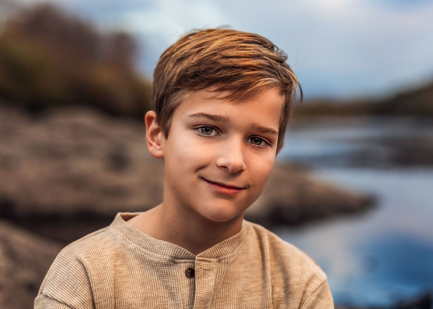 A young boy with brown hair smiles softly outdoors near a rocky lake under a cloudy sky.
